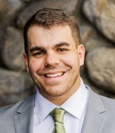 A young man is smiling and dressed in a blue suit with a light blue shirt and a striped tie. He is standing outdoors on a pathway with trees and greenery in the background.