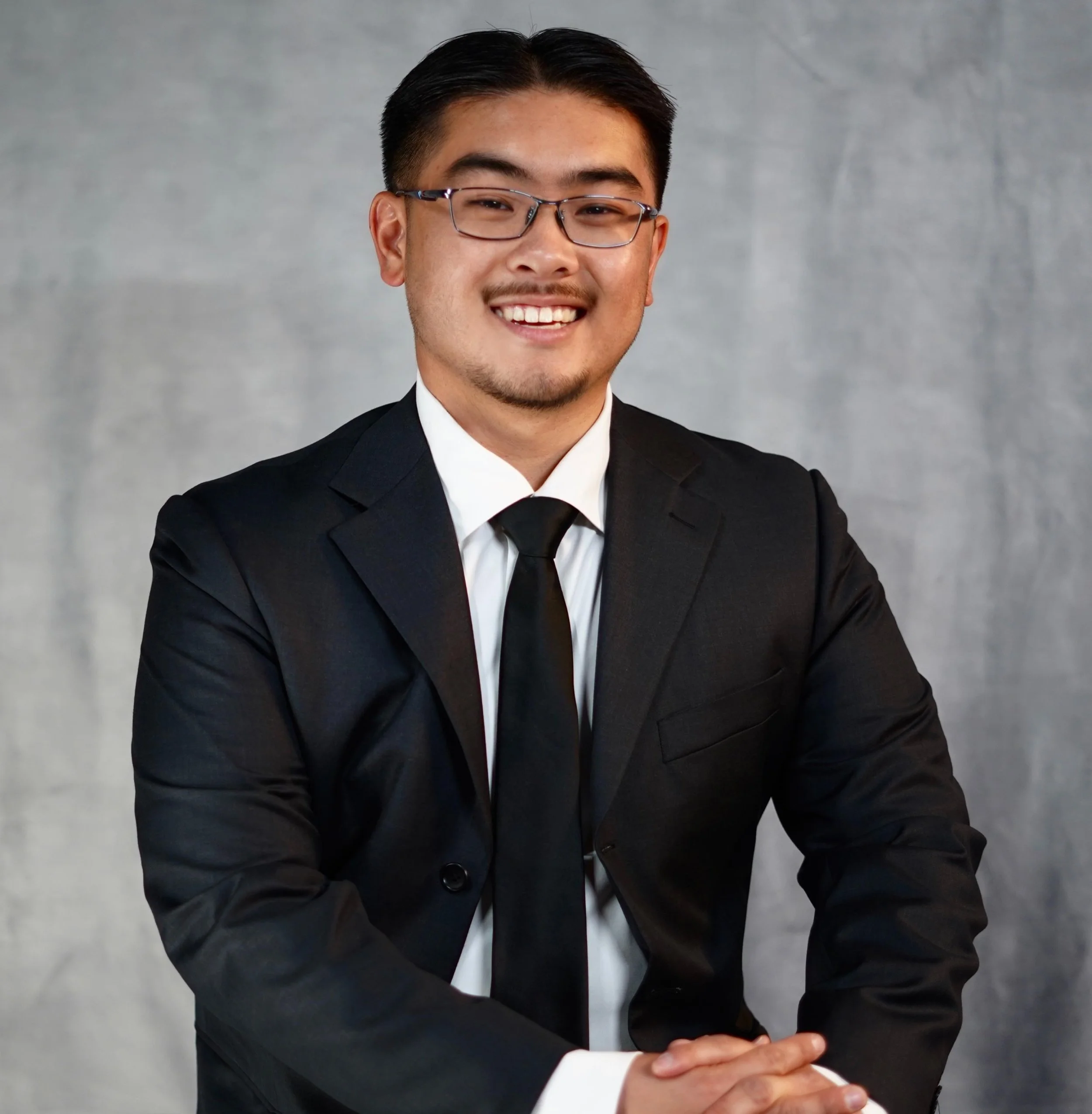 A young man in a black suit, white shirt, and black tie, smiling and posing with folded hands against a gray background.