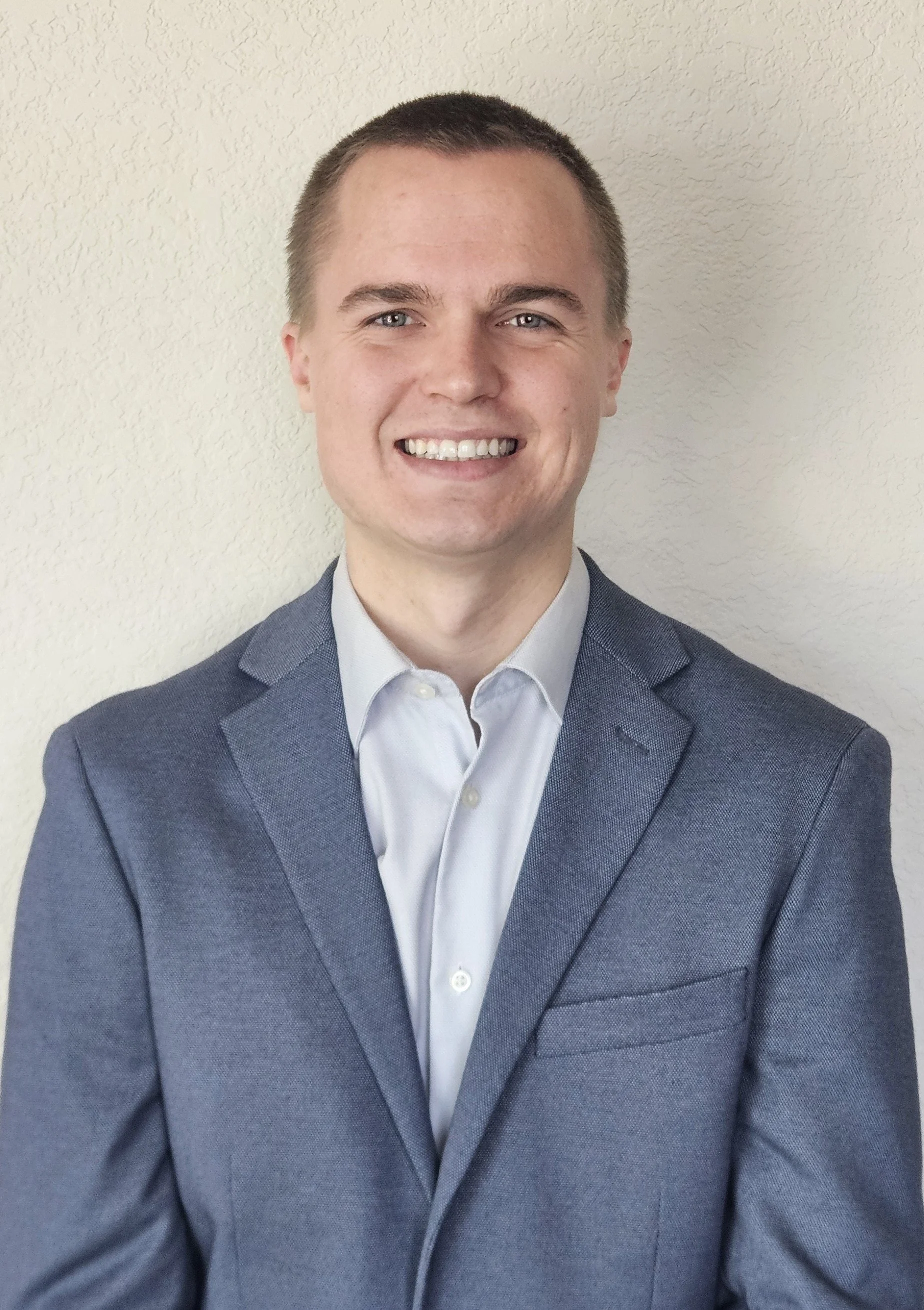 A young man with short brown hair, light skin, and blue eyes wearing a dark blue suit jacket and a white dress shirt, smiling and standing against a plain, off-white textured wall.