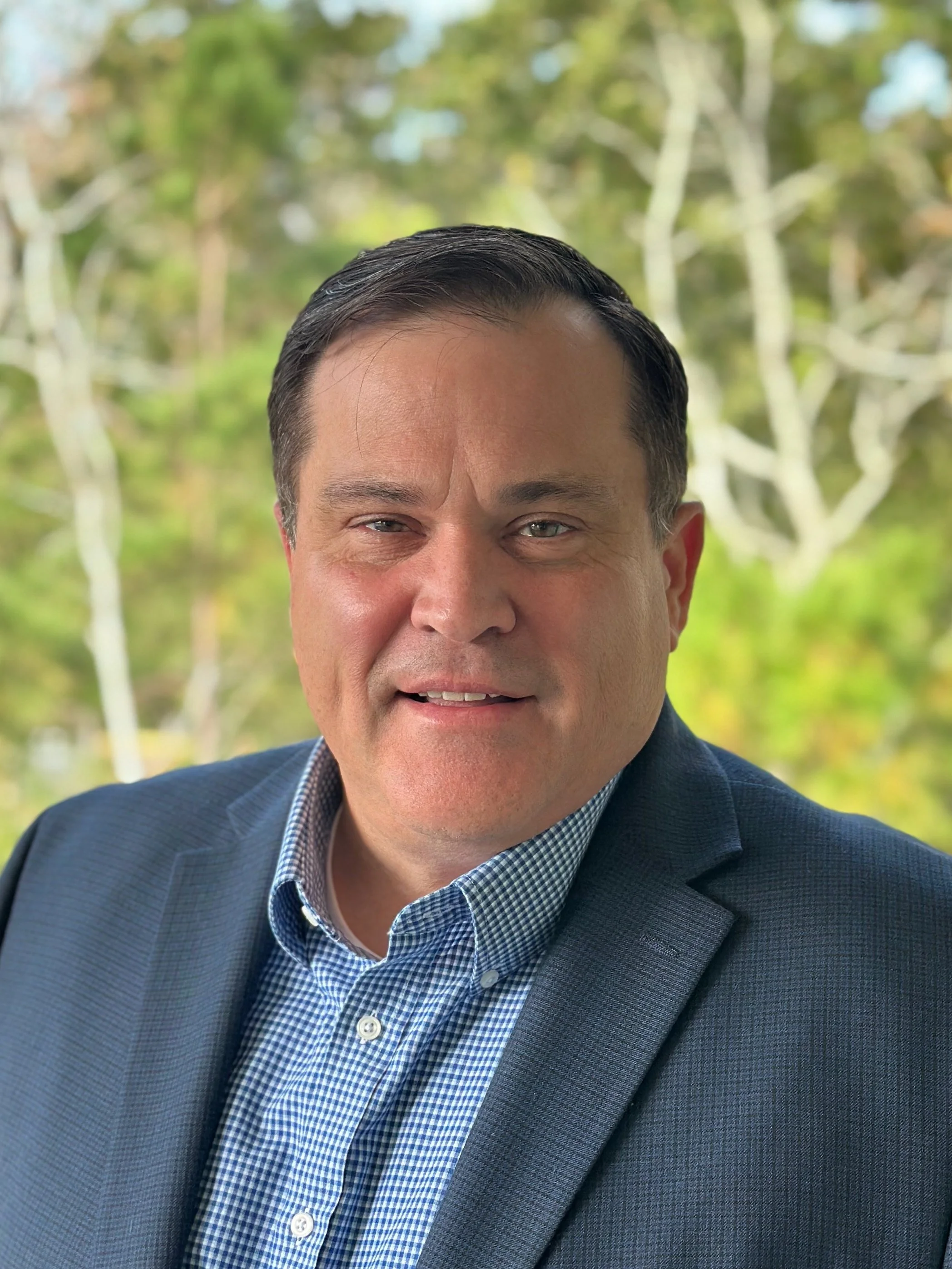 Portrait of a man in a suit with a blurred green outdoor background.