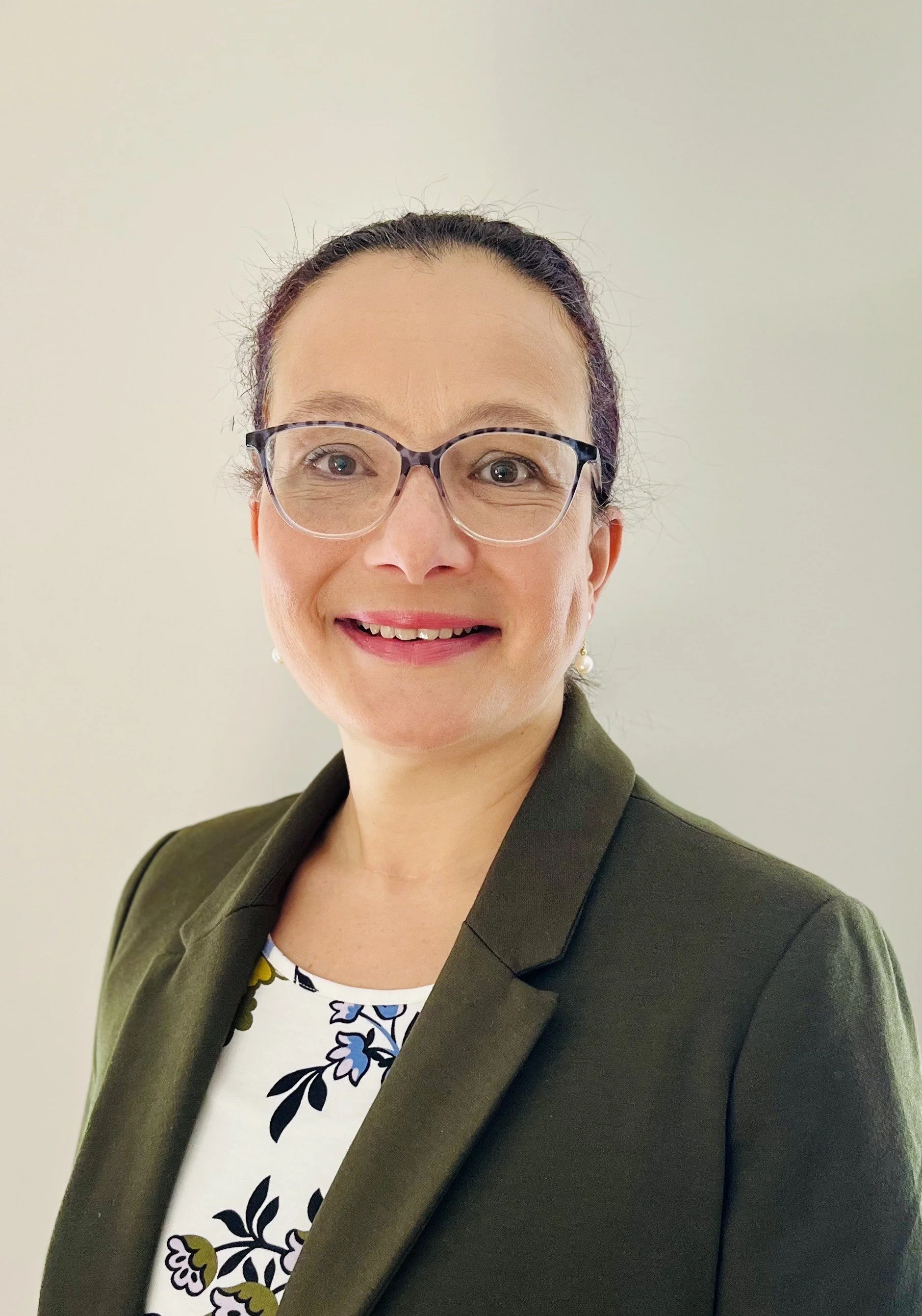 A woman with glasses and earrings smiling, wearing a dark green blazer and a floral blouse, standing against a plain light-colored wall.