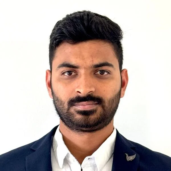 A young man with dark hair and a beard, wearing a navy blue suit jacket and a white shirt, standing against a light-colored background.