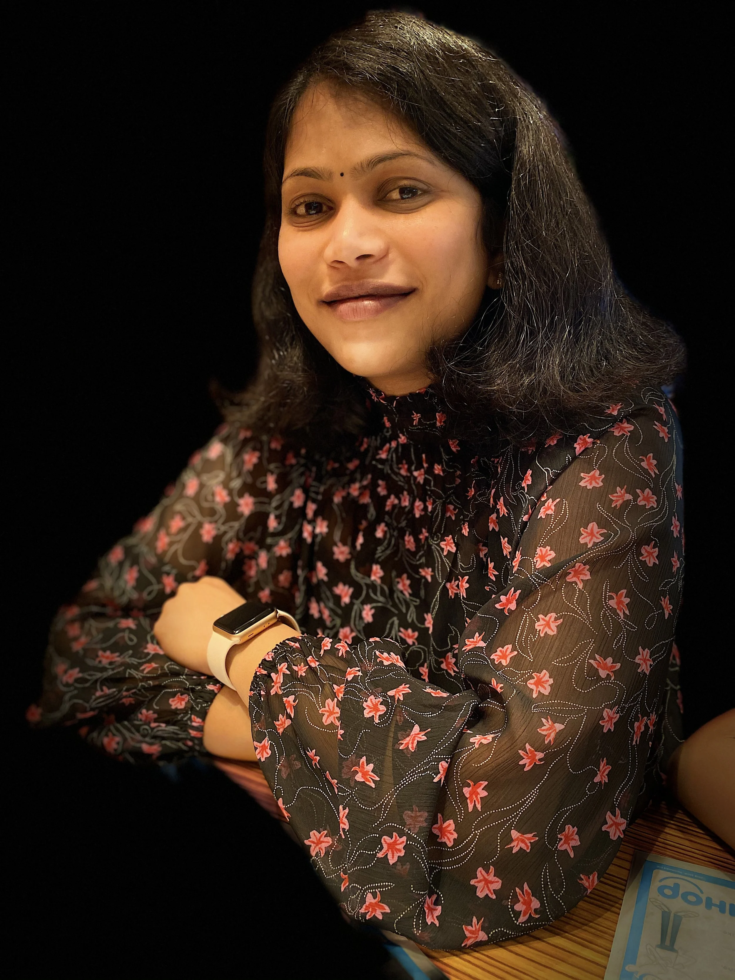 A woman with dark, shoulder-length hair, wearing a black blouse with red and pink floral patterns, sitting with arms crossed on a wooden table and smiling.
