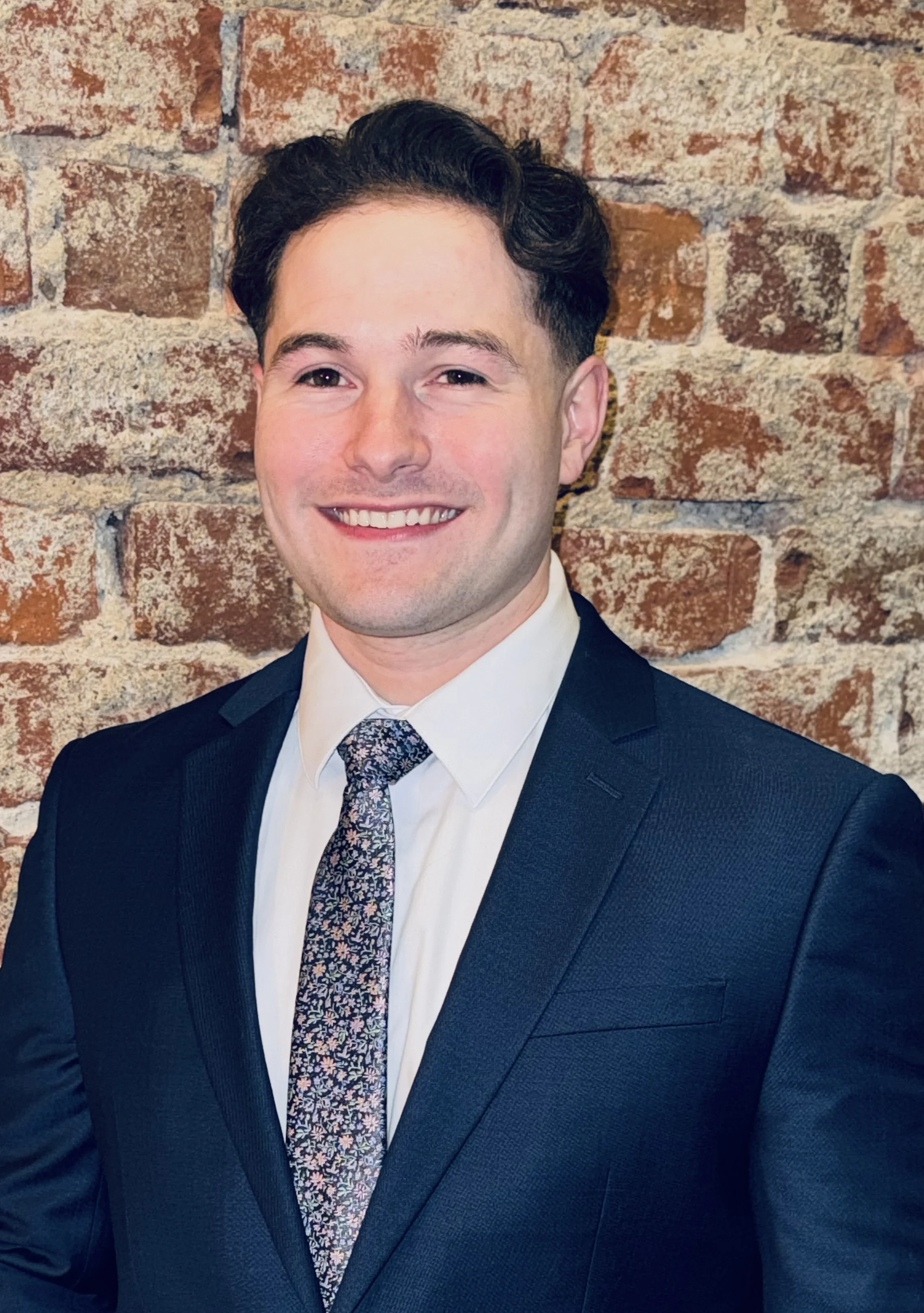 A young man in a dark suit, white shirt, and patterned tie smiling in front of a brick wall.
