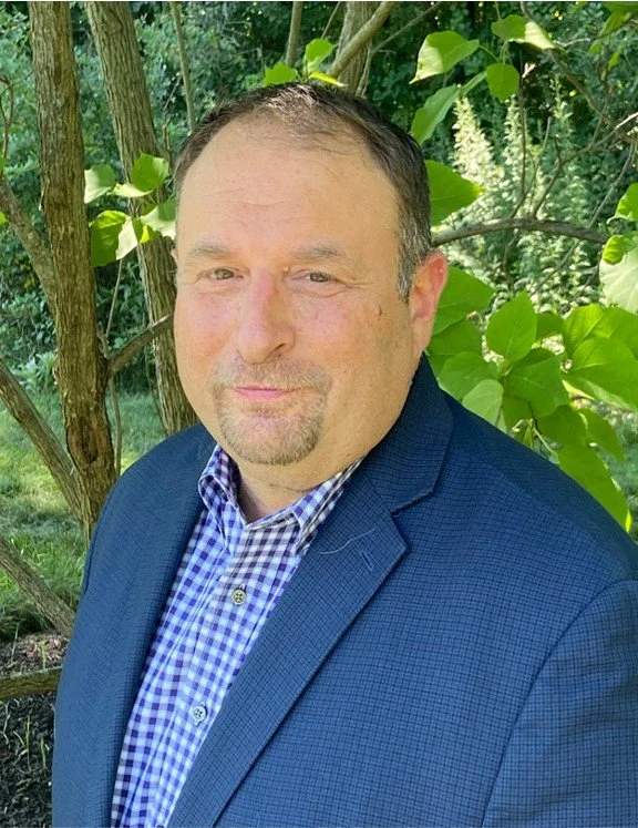 A man wearing a blue checkered shirt and navy blazer standing outdoors with green foliage and trees in the background.