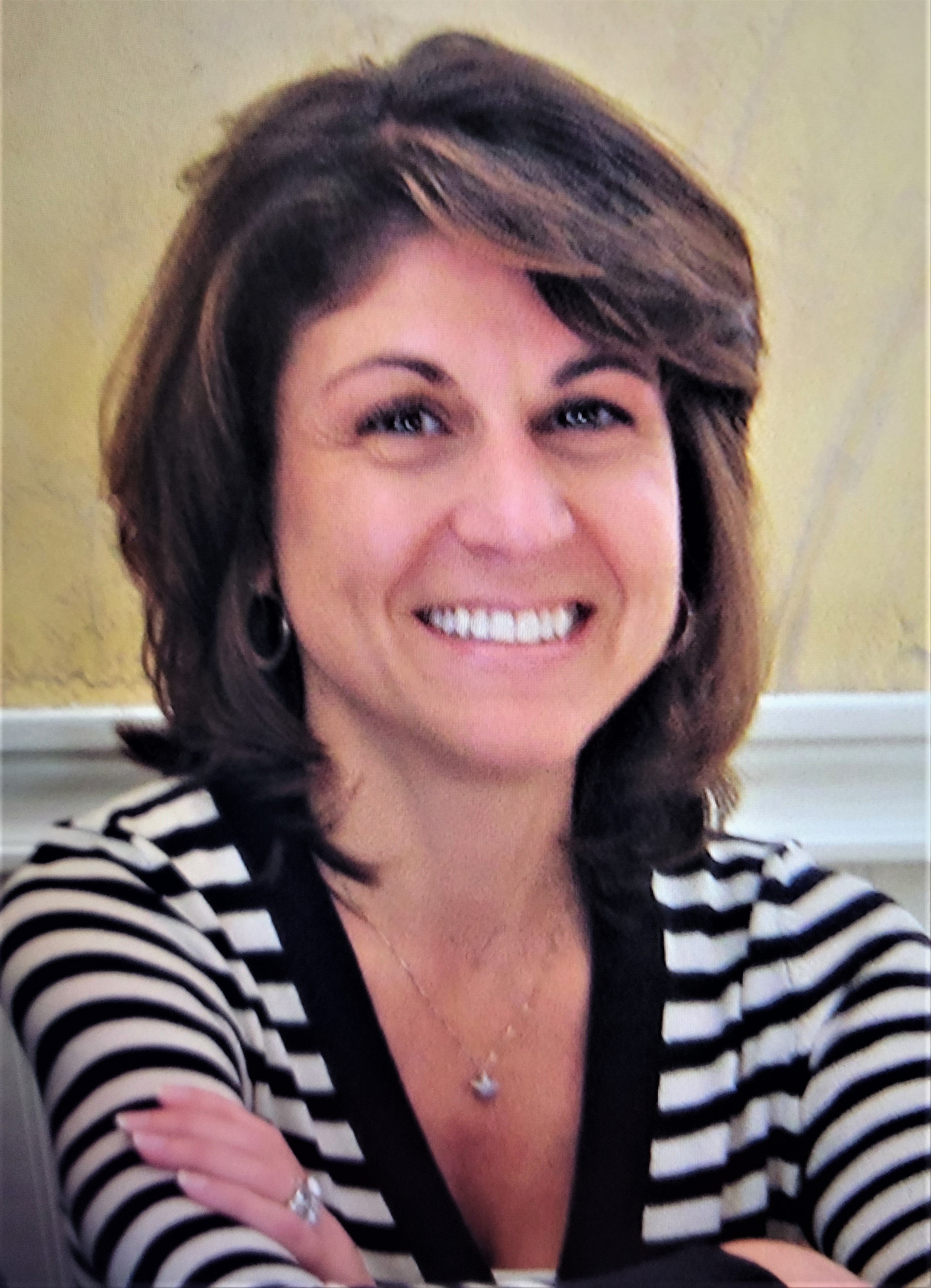 A woman with shoulder-length brown hair, smiling broadly, wearing a black and white striped top, a necklace, earrings, and a ring, sitting with arms crossed in front of a yellow wall.