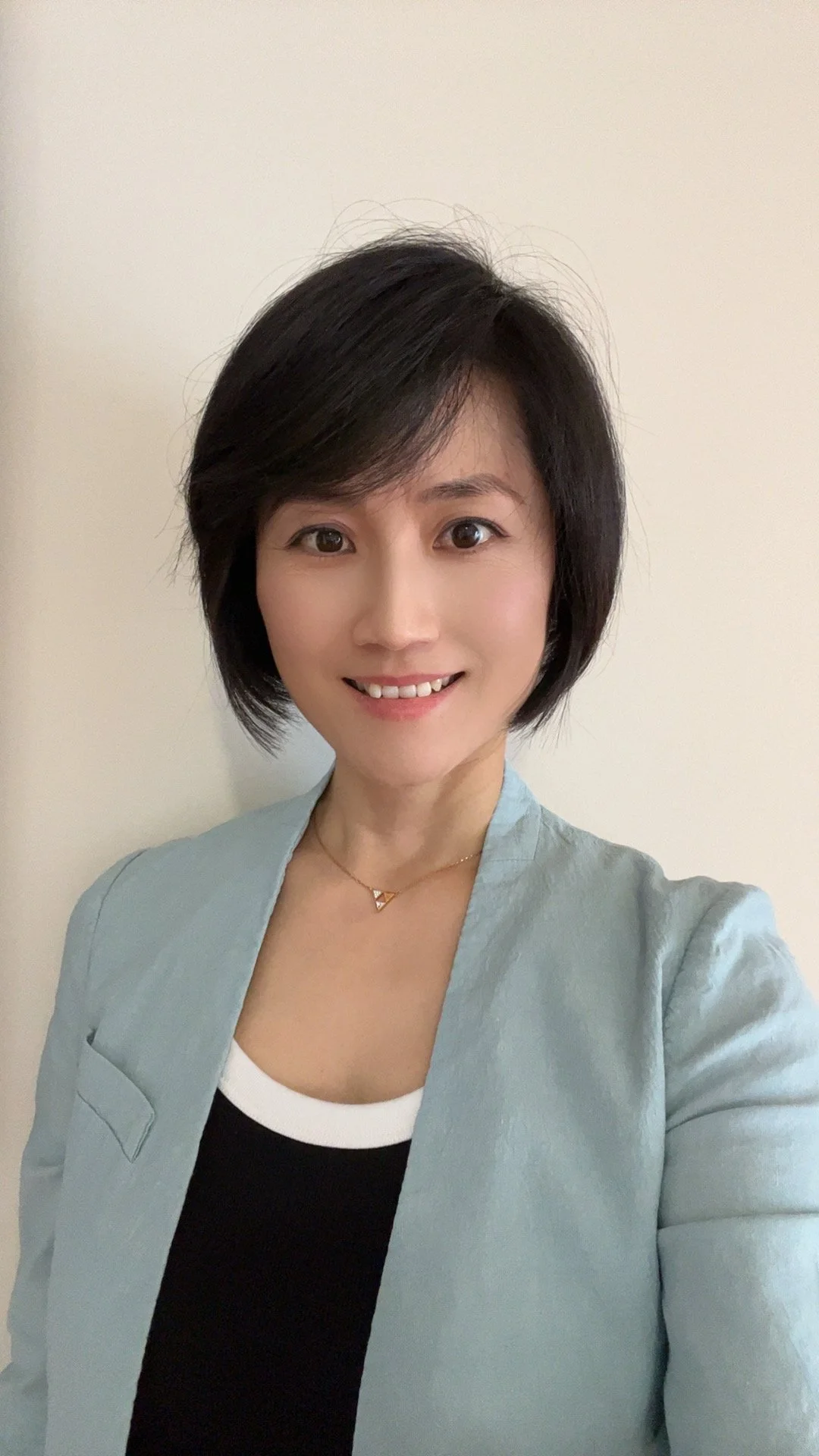 A woman with short black hair, wearing a black blazer and light makeup, taking a selfie in front of a plain white wall.