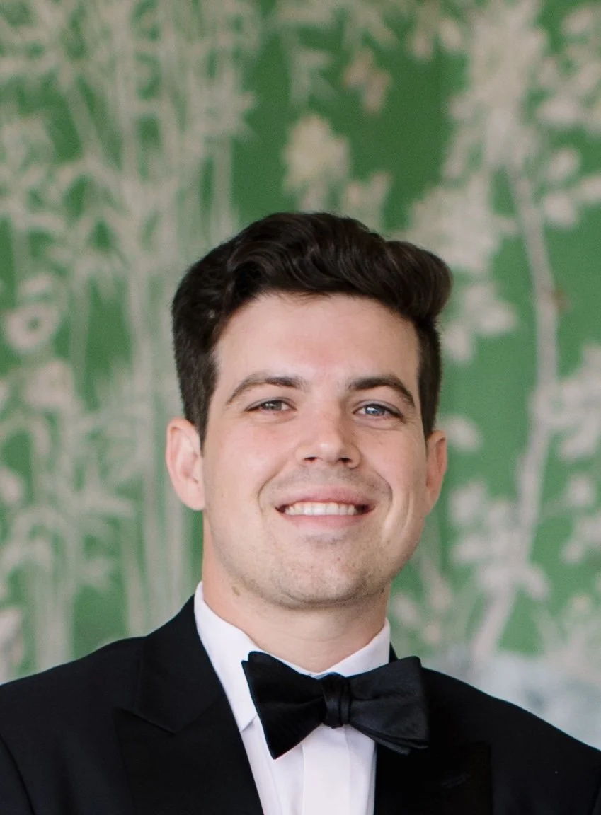Portrait of a smiling young man with brown hair and blue eyes, wearing a black collared shirt, standing against a plain light-colored wall.