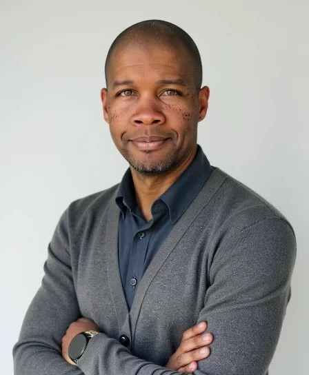 A man with short hair, wearing a dark blue shirt and gray sweater, standing with arms crossed against a plain light gray background.