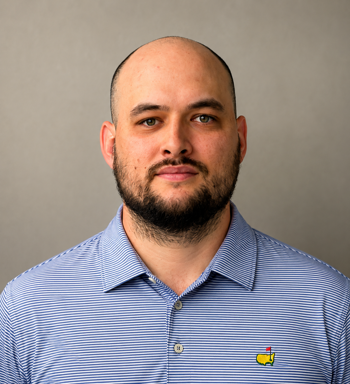 A smiling man with dark hair and a beard, wearing a navy blazer and a checkered shirt, against a plain white background.