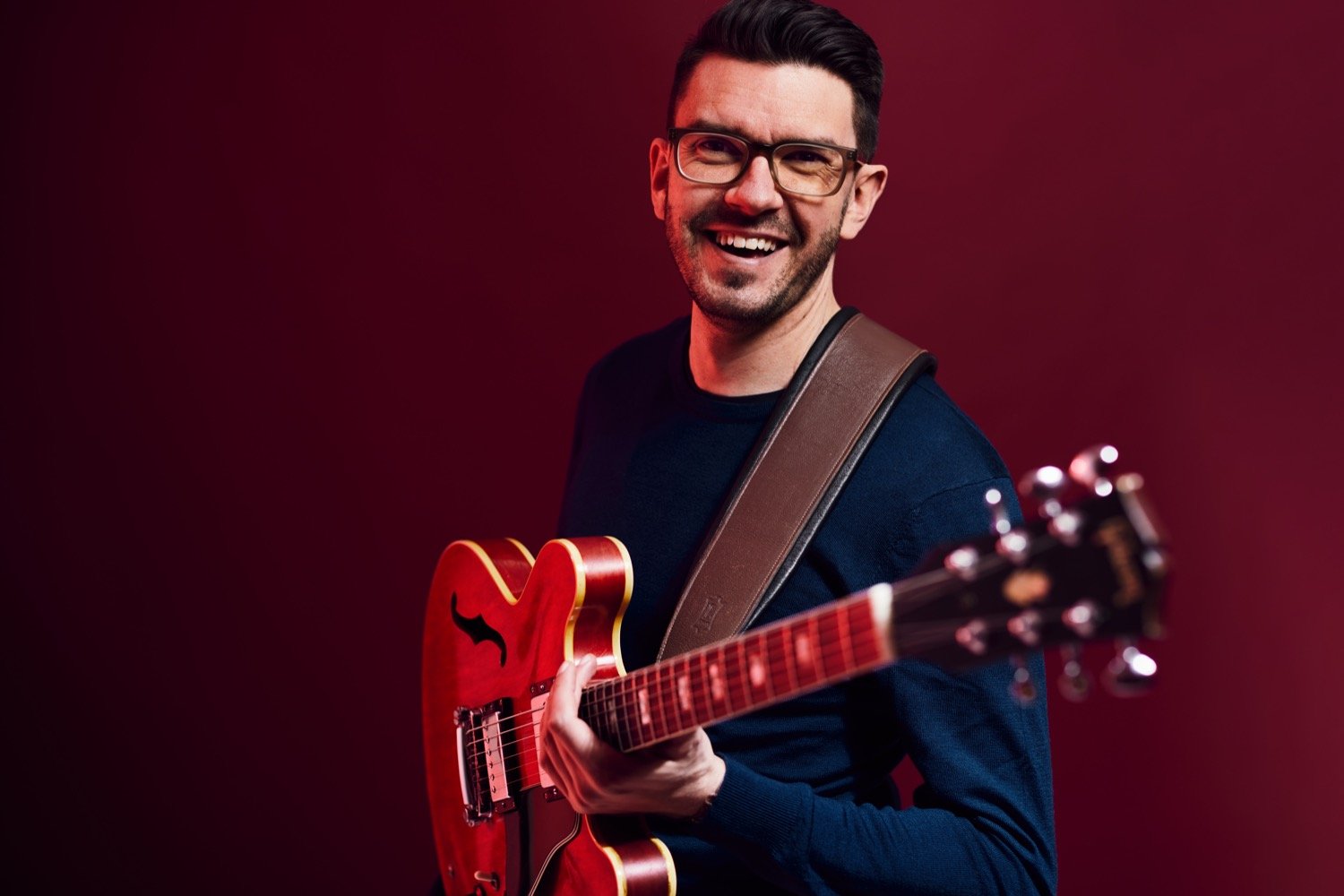 landscape-studio-steve-banks-loose-side-on-laughing-holding-guitar-red-background-profile-image-hero-colour.jpg