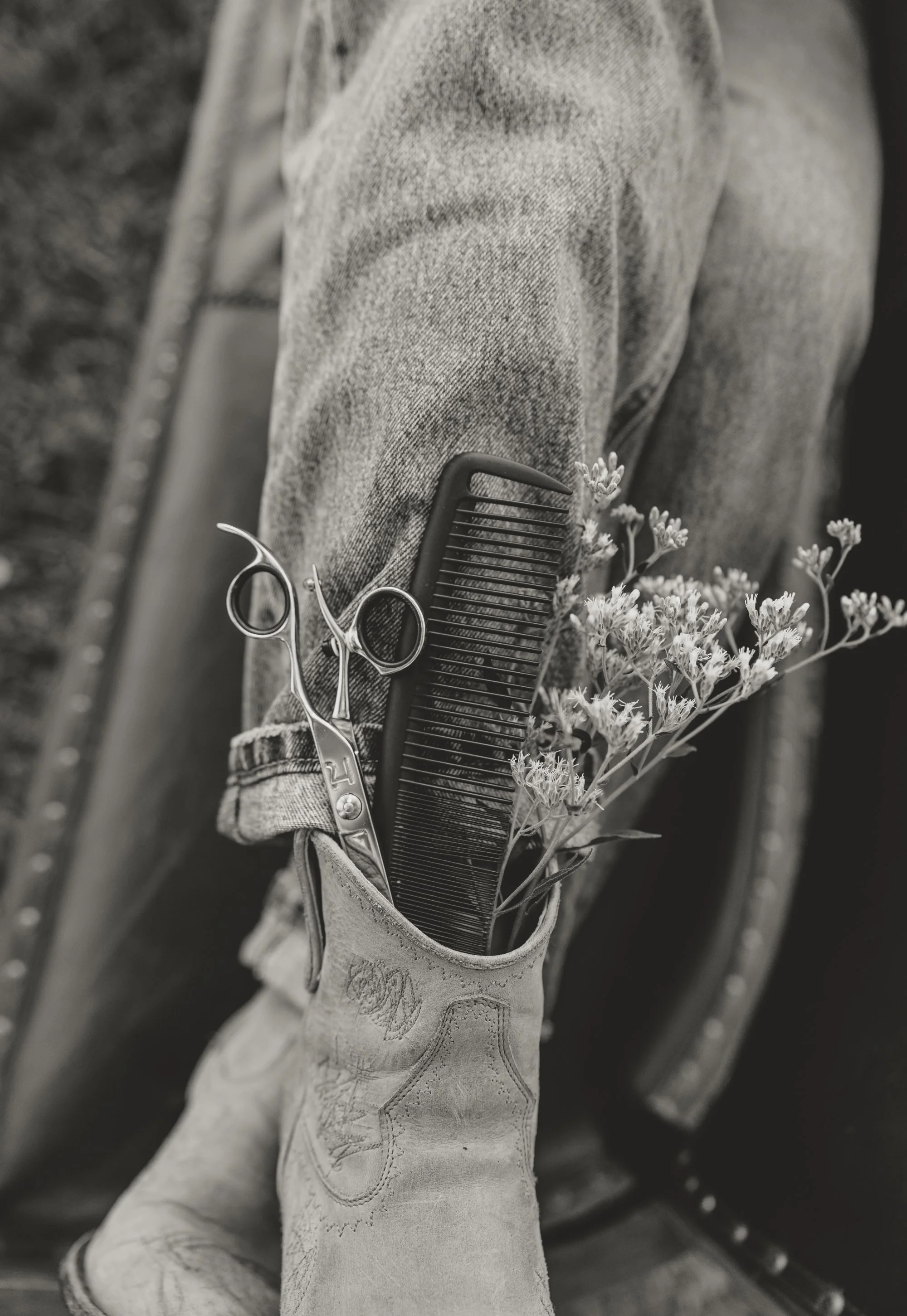 Close-up of a work boot with a small branch of flowers tucked into its opening, with grooming scissors and a comb resting inside the boot, all resting against a denim fabric background.