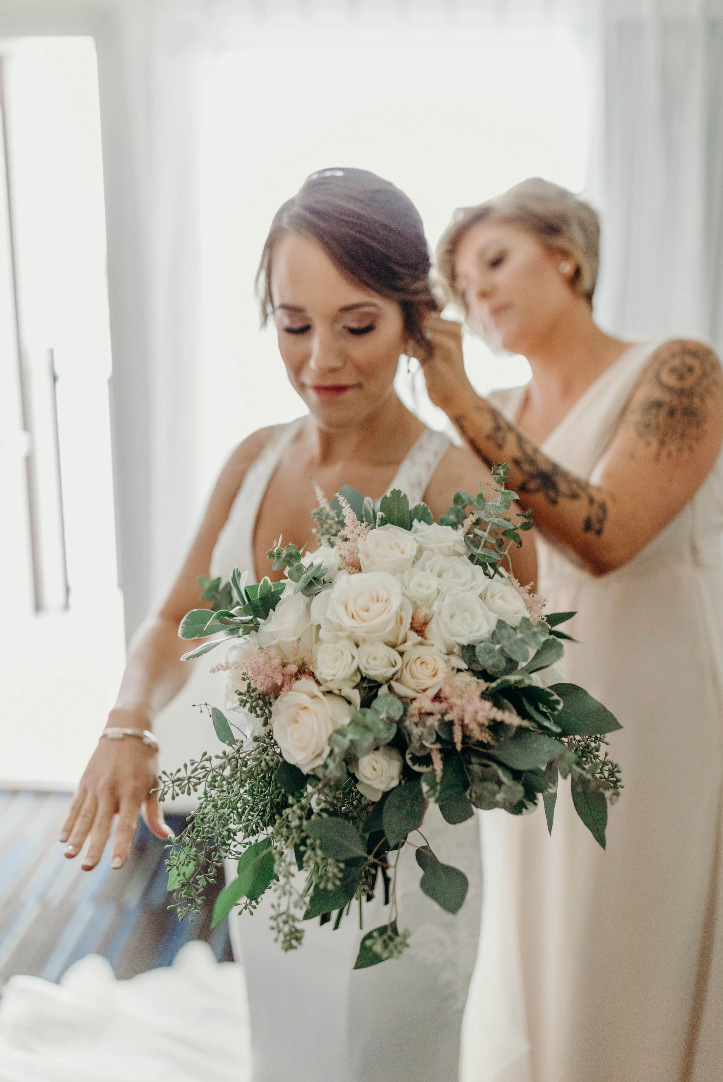 A bride holding a large bouquet of white and blush roses with greenery, standing in a softly lit room, while a woman helps her with her dress or hair.