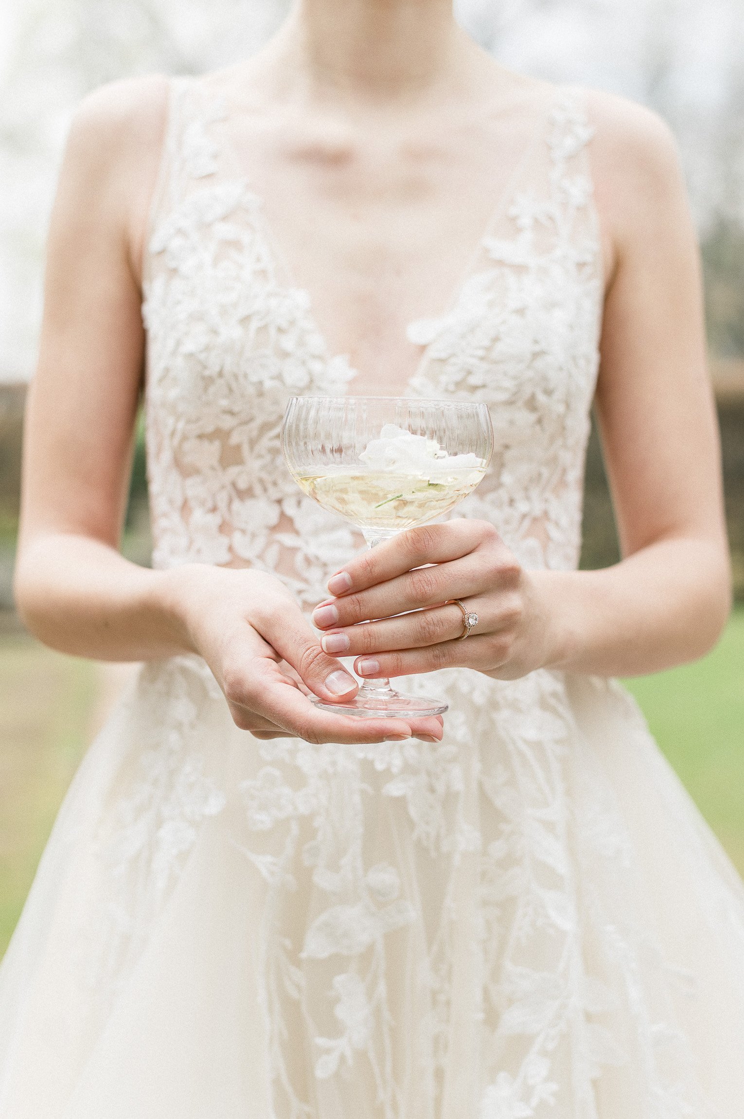 A woman in a white lace wedding dress holding a champagne glass with a floral decoration inside, outdoors with blurred background.