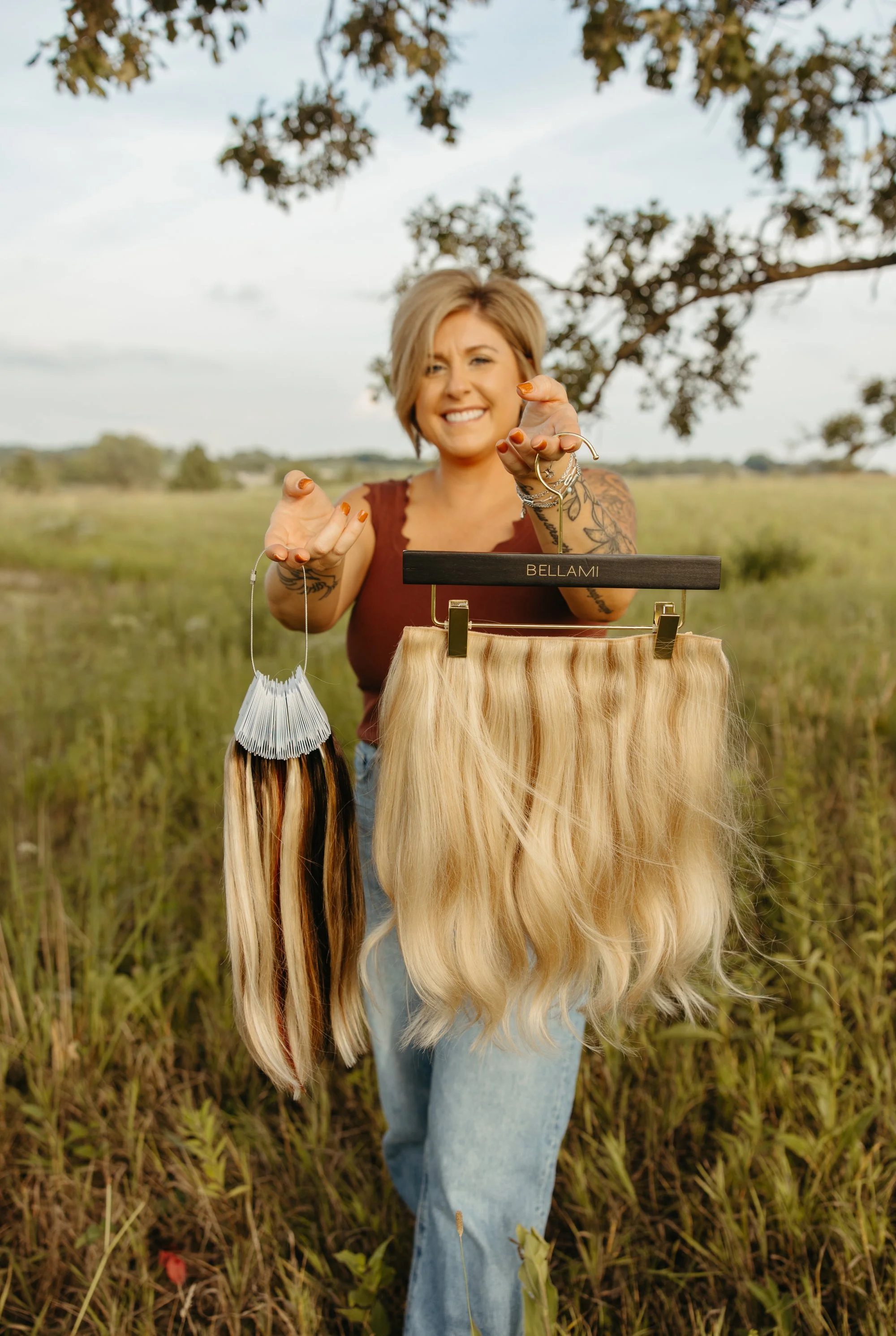 Woman smiling outdoors holding a wig on a hanger labeled 'BELLAMI' in a grassy field with trees in the background.
