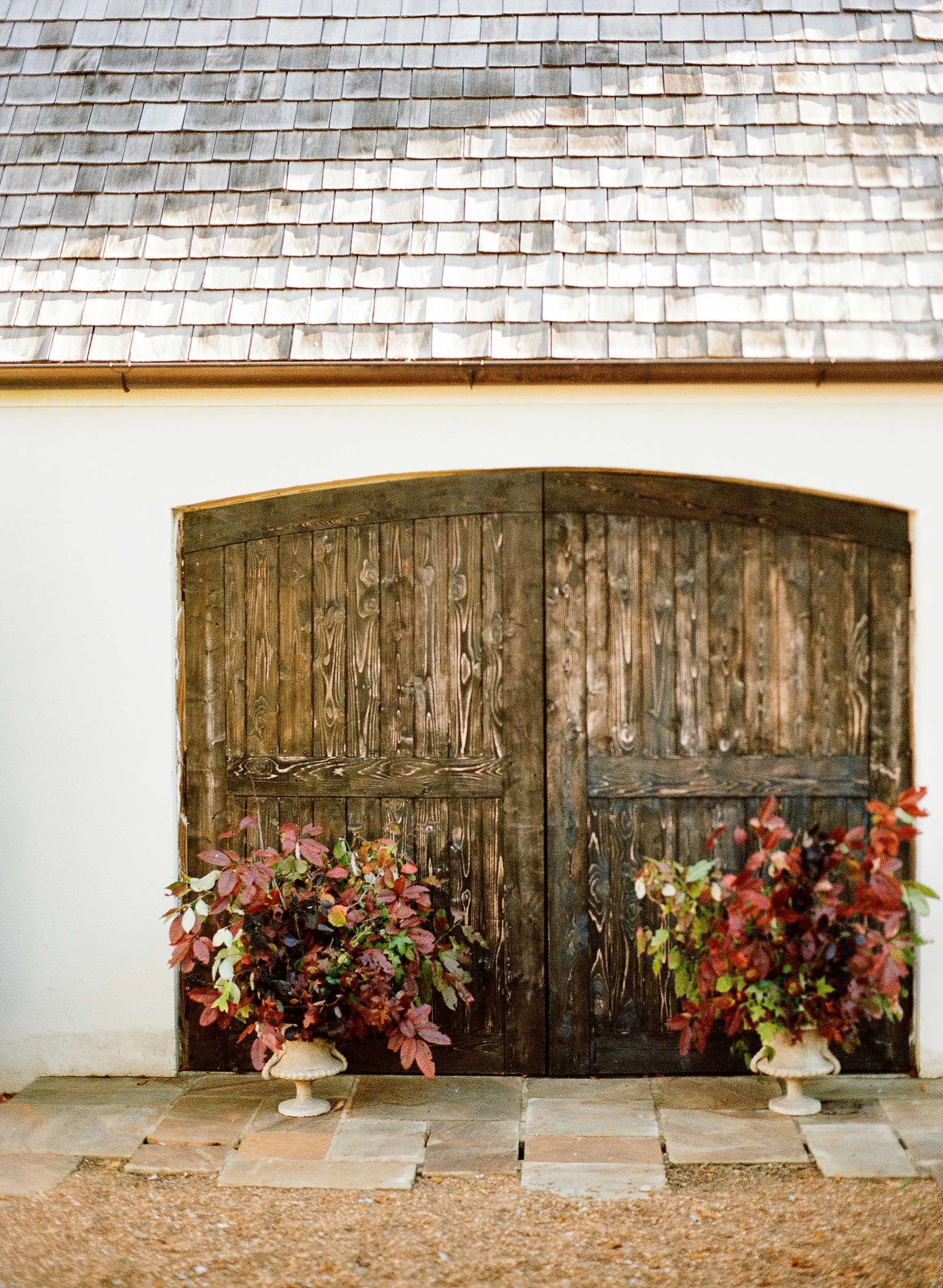 A wooden set of double doors with an arched top, flanked by two potted bushes with reddish leaves. The doors and bushes are positioned in front of a white wall, with a tiled roof above.