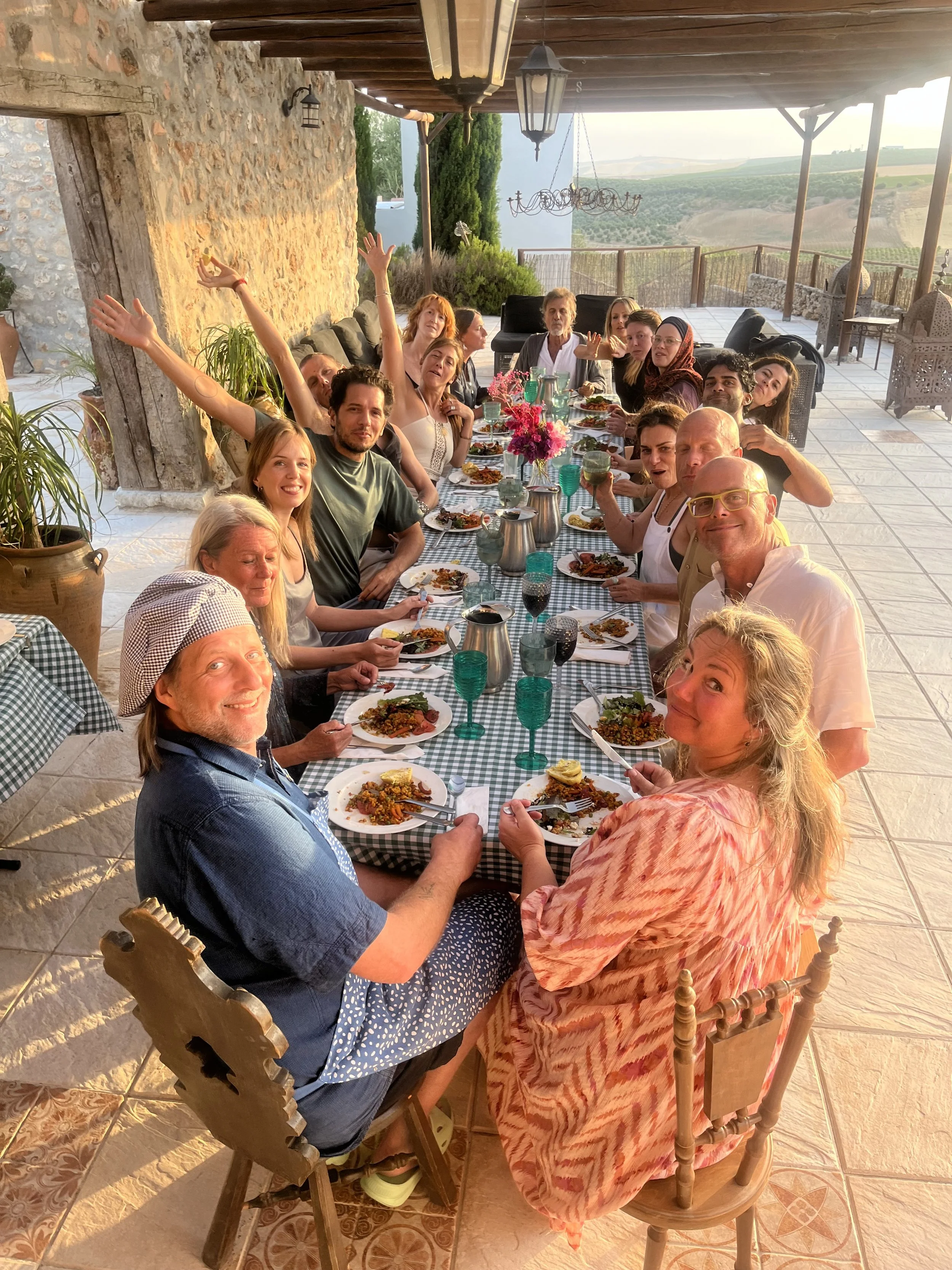 A large group of people sitting at a long outdoor dining table, enjoying a meal together on a patio with scenic countryside in the background. The table is decorated with a pink bouquet of flowers, and the setting is illuminated by warm sunset lighting.