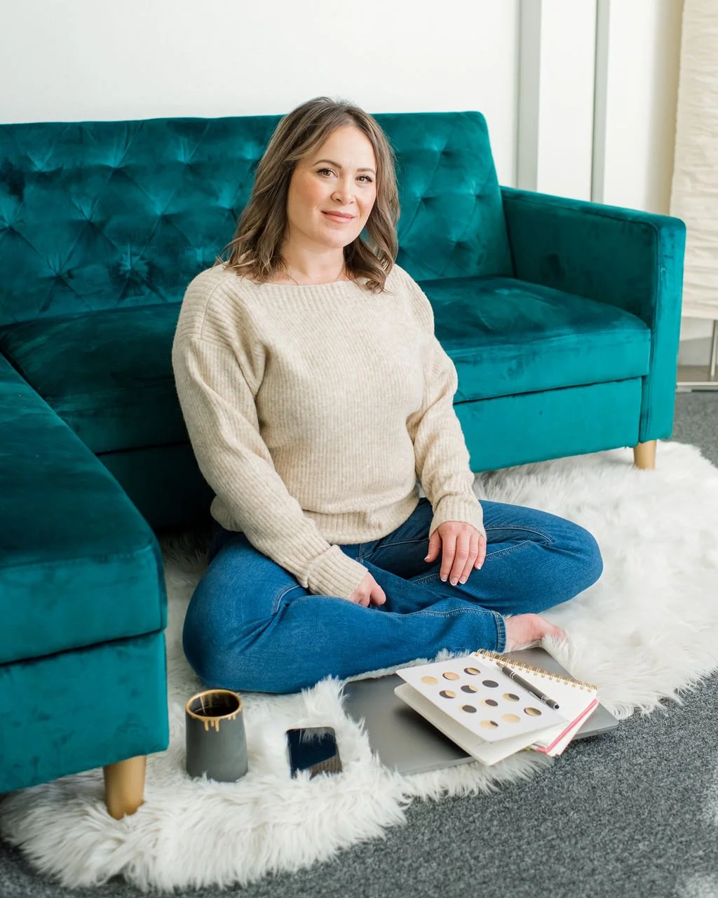 A woman sitting cross-legged on a white fluffy rug in front of a teal velvet sofa, with a notebook, a black coffee mug, a smartphone, and a brochure or catalog beside her.