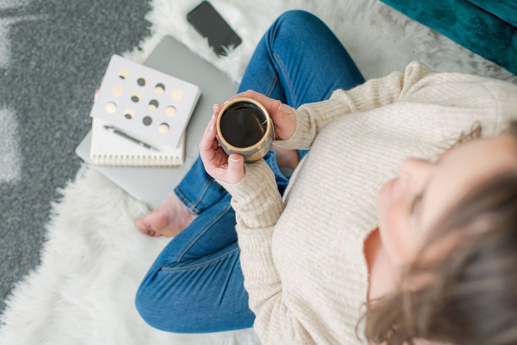 Person sitting on a white fluffy rug, holding a coffee cup, with notebooks, a pen, and a black smartphone nearby.