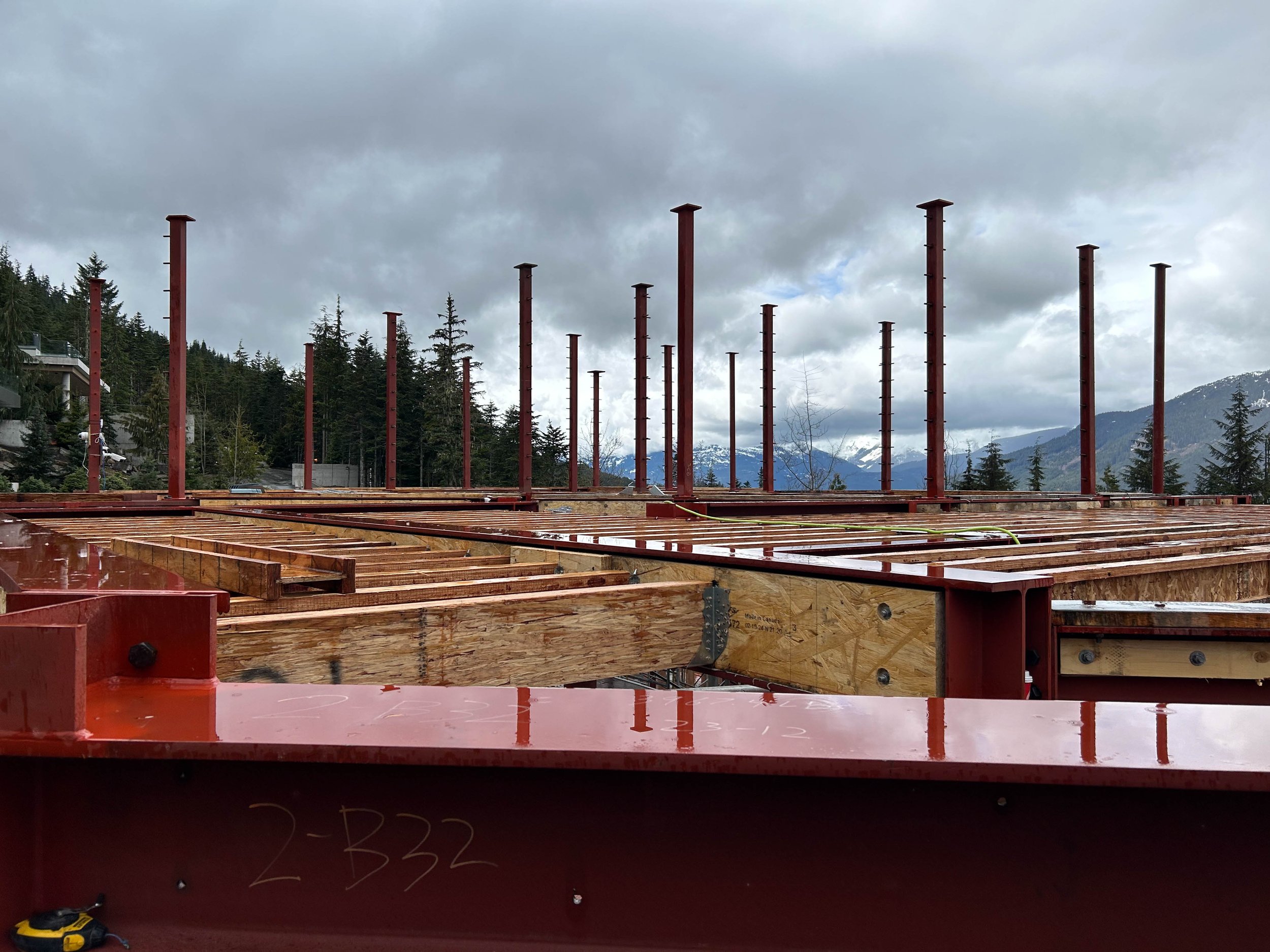Construction site with wooden beams and red steel supports, set against a backdrop of forested mountains and cloudy sky.