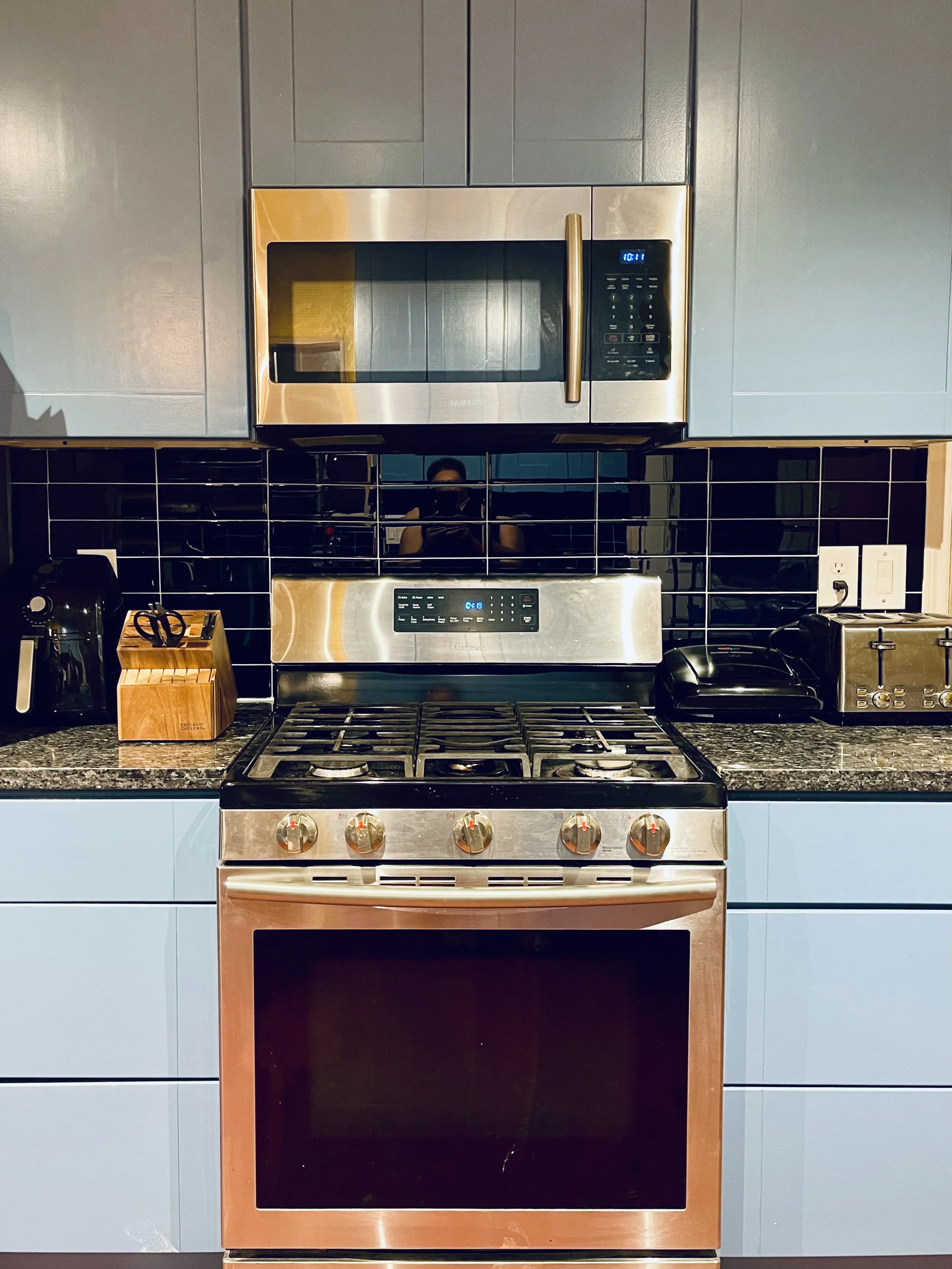 Modern kitchen with stainless steel microwave and oven, black backsplash, blue cabinets, granite countertop, toaster, kettle, and utensil holder.