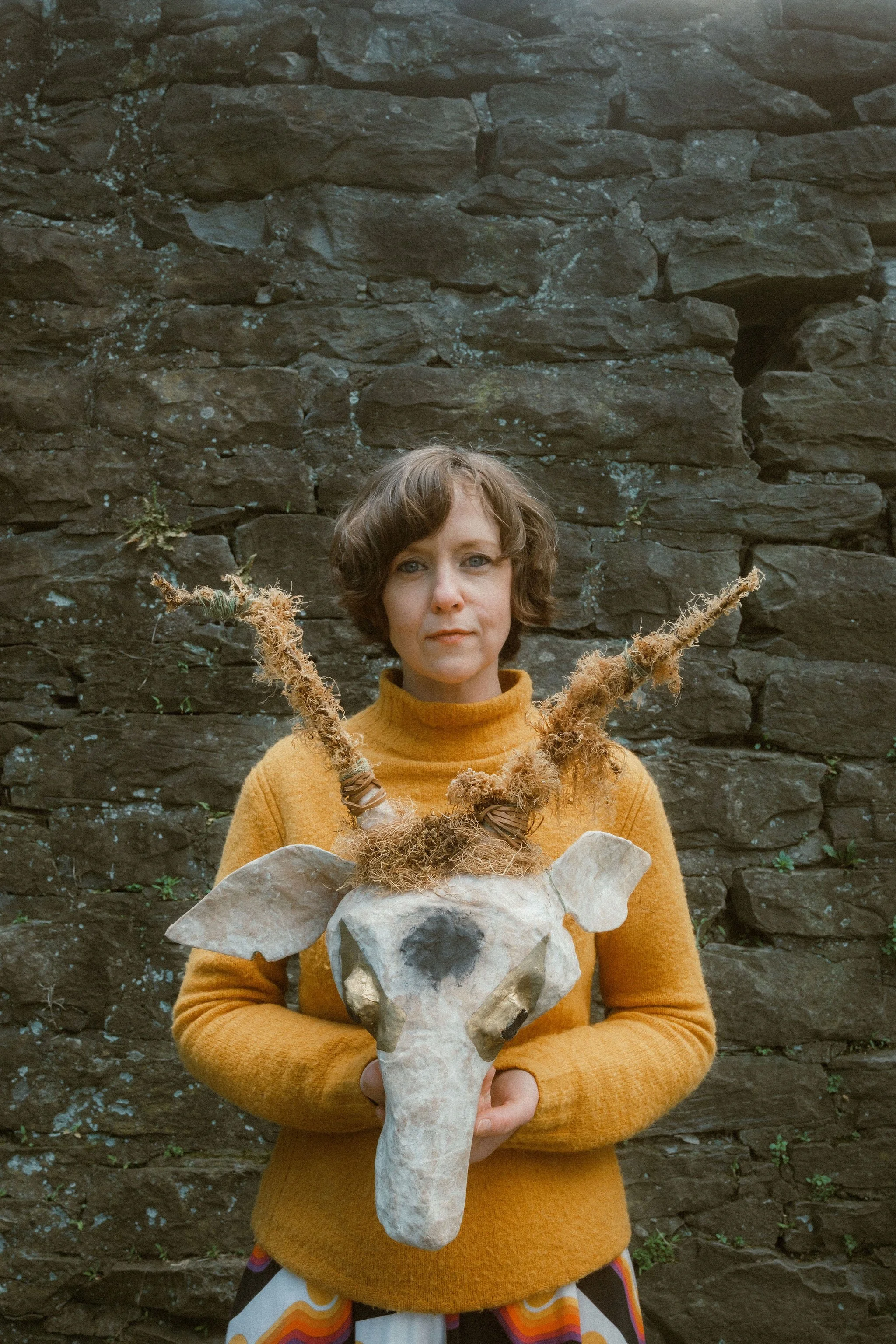 A white woman with short brown hair is wearing a yellow sweater and smiling while she looks at the camera. She is holding a stag mask made out of papier mache, branches, and dried brown moss.
