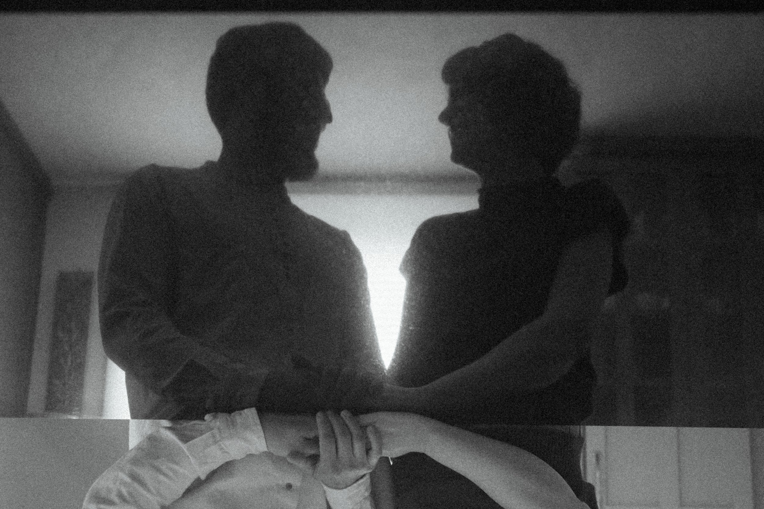A black and gray image of Yigit and Kait, reflected in a shiny black surface. They are holding hands which appear to be upside in the very bottom of the frame, due to the tricky reflection.