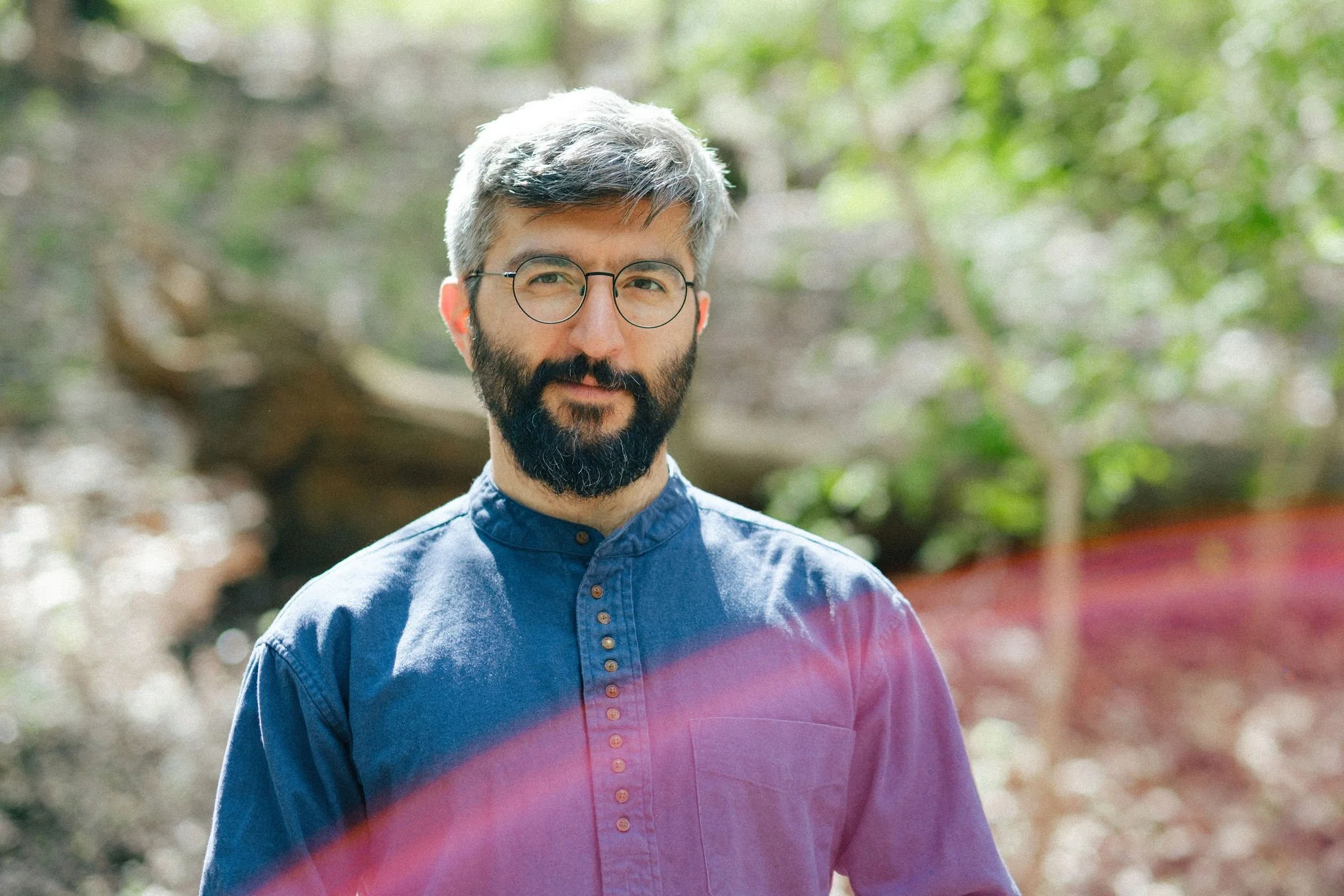 A man with a brown complexion and short gray-and-black hair is wearing black, round, wire-rimmed glasses and a long-sleeved, blue, linen shirt. He is standing in the woods and smiling at the camera. The sun is peaking through the tree canopy onto him