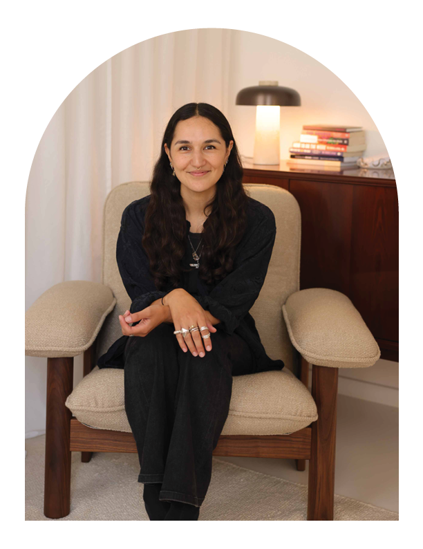 Hue Therapy | Sabrina | A woman with long dark hair sitting on a beige armchair in a cozy room, smiling at the camera. There is a wooden side table with stacked books and a lamp behind her.