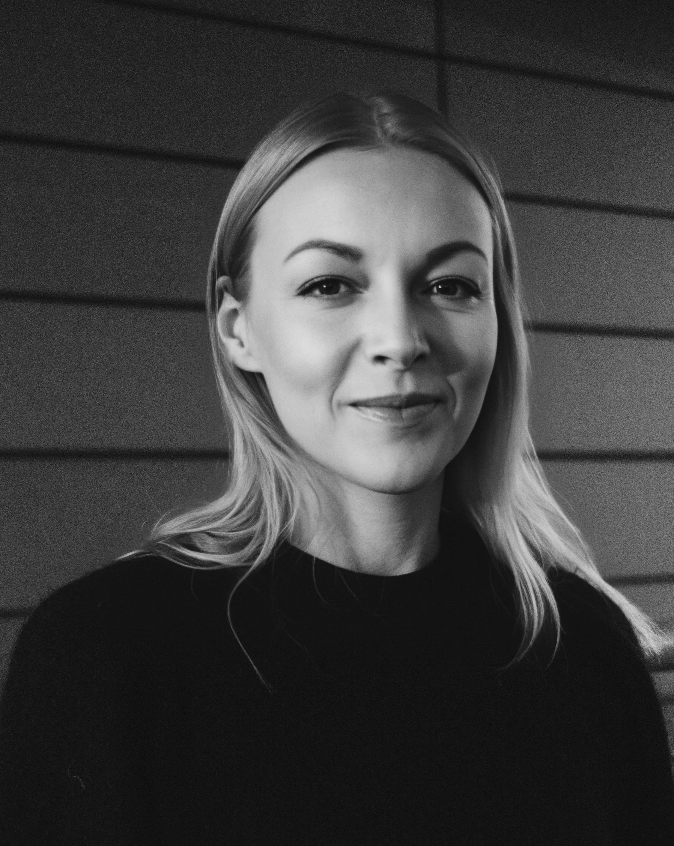 Black and white portrait of a woman with shoulder-length hair, smiling softly, standing in front of a wooden-paneled wall.