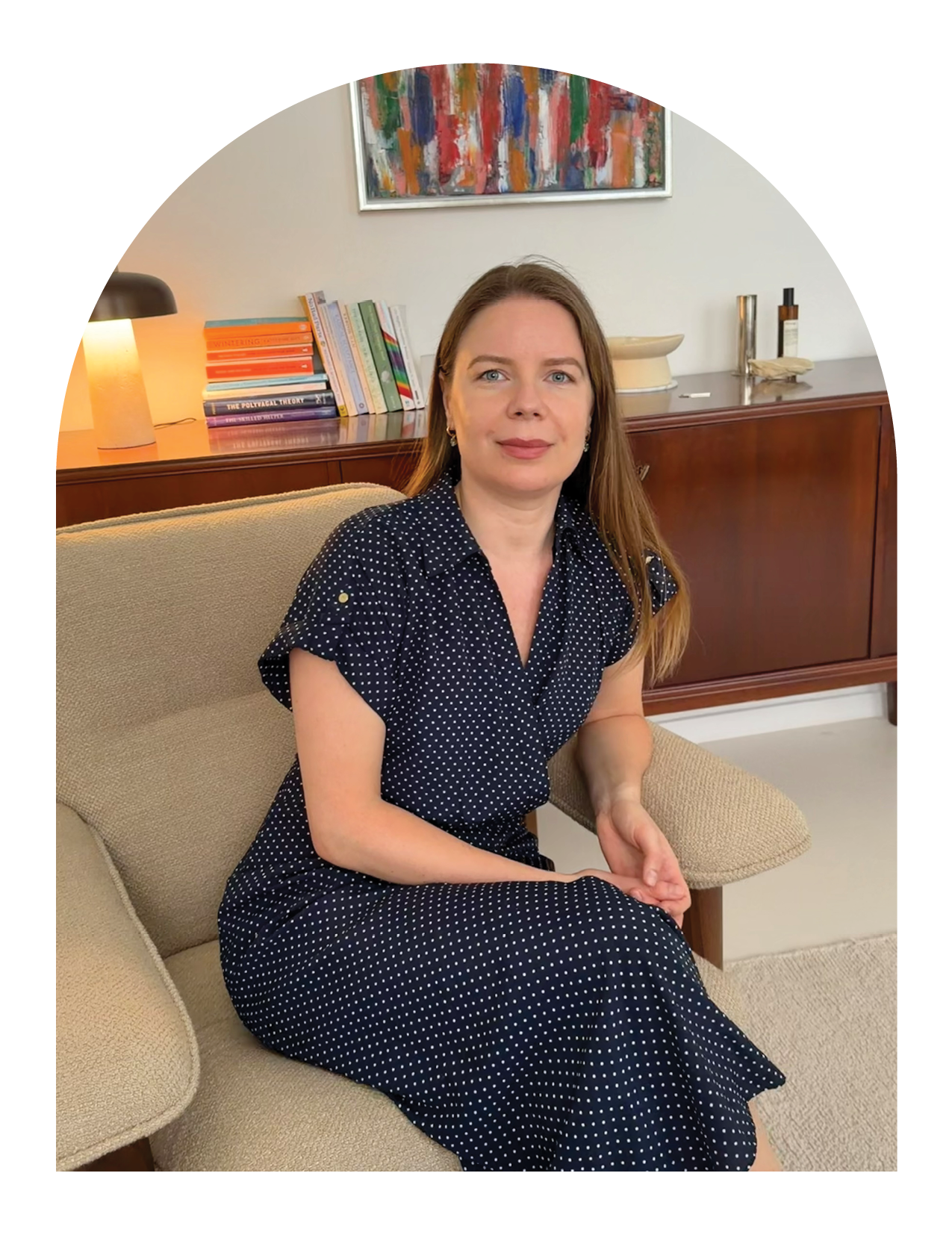Hue Therapy | Nelda | A woman with long dark hair sitting on a beige armchair in a cozy room, smiling at the camera. There is a wooden side table with stacked books and a lamp behind her.