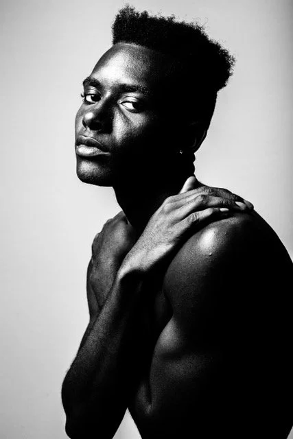 A black and white portrait of a shirtless person with short curly hair, posing with one hand on their shoulder and looking at the camera.