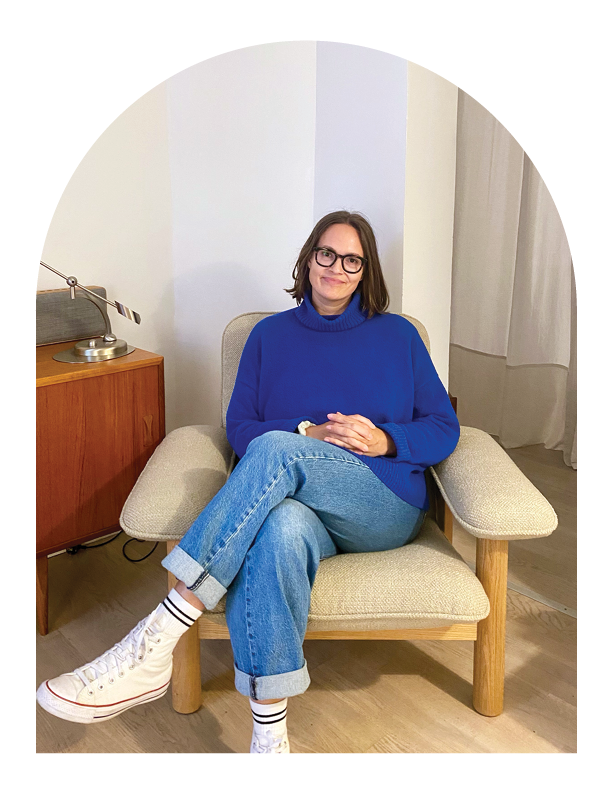 Hue Therapy | Marta | A woman with long dark hair sitting on a beige armchair in a cozy room, smiling at the camera. There is a wooden side table with stacked books and a lamp behind her.