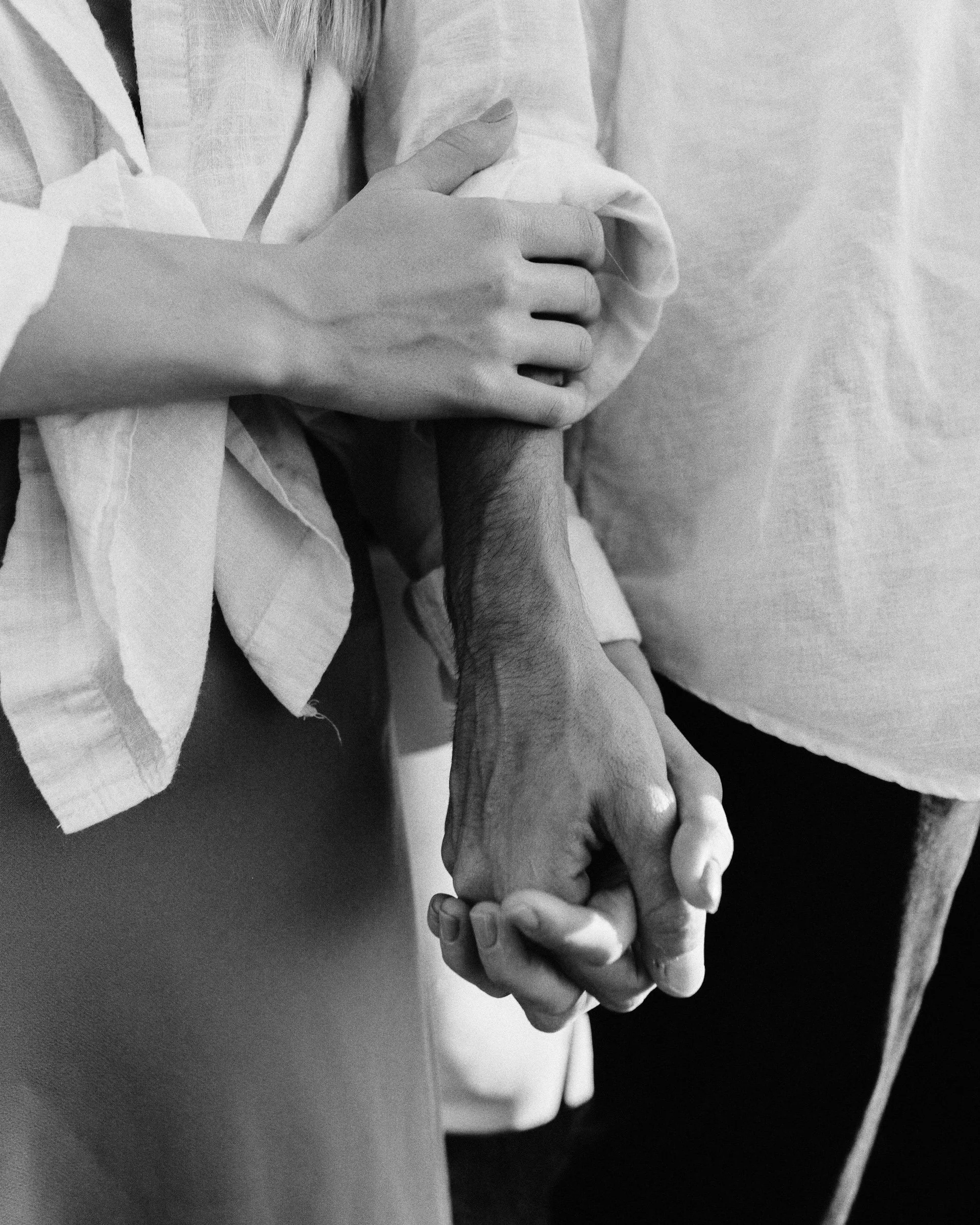 Close-up of two people holding hands, one with a lighter hand and arm and the other with a darker, hairier arm, in black and white
