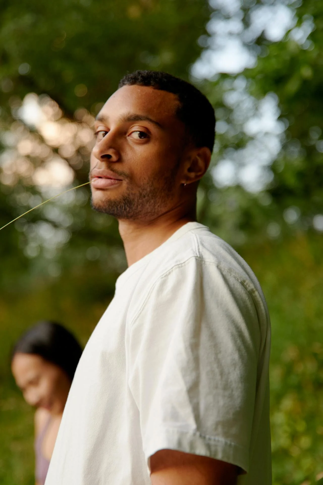 A young man with short dark hair and facial hair looking at the camera, while a woman stands in the background outdoors surrounded by green trees.