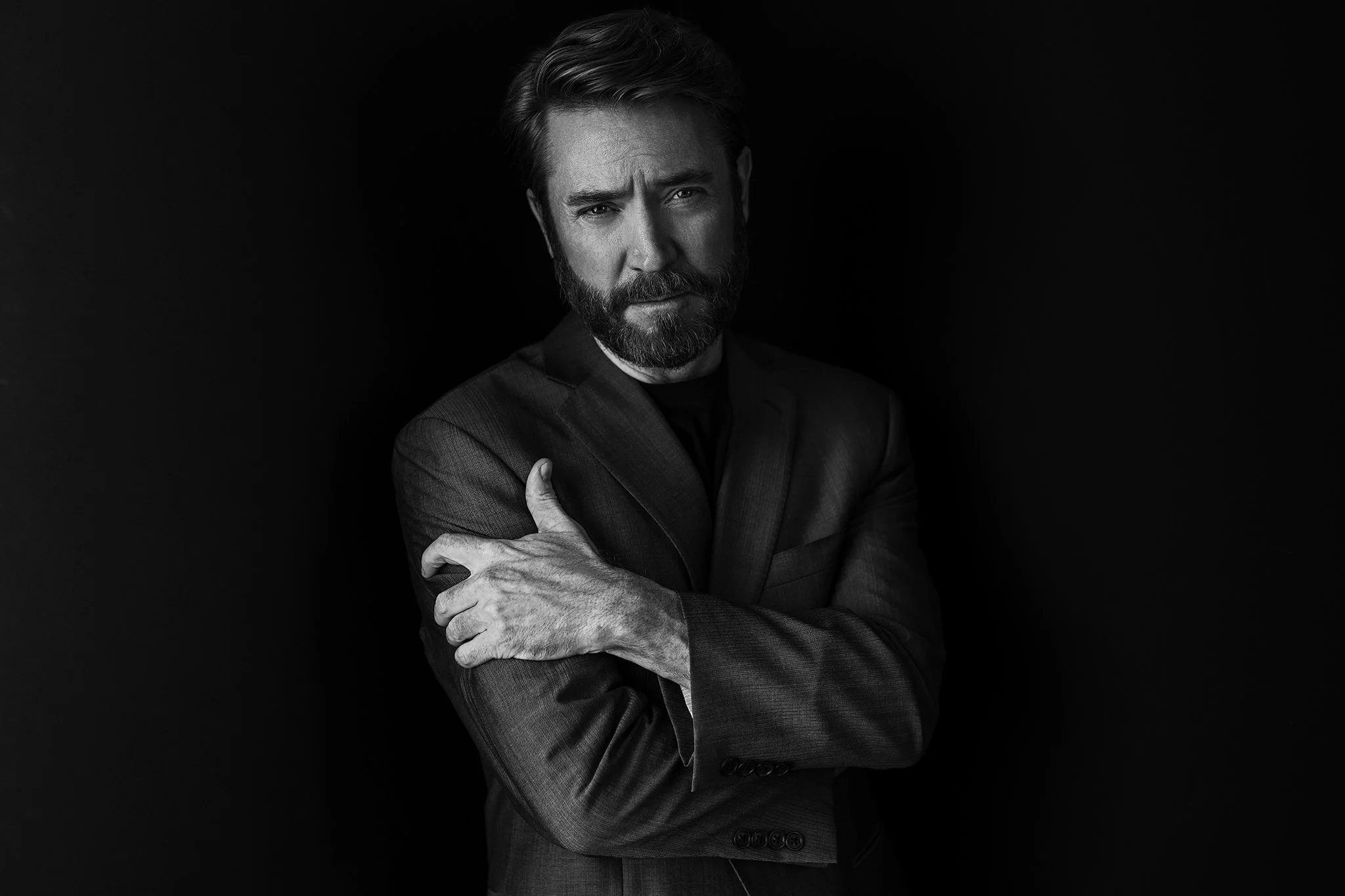 Black and white portrait of a man with a beard and styled hair, wearing a suit, standing against a dark background with arms crossed.