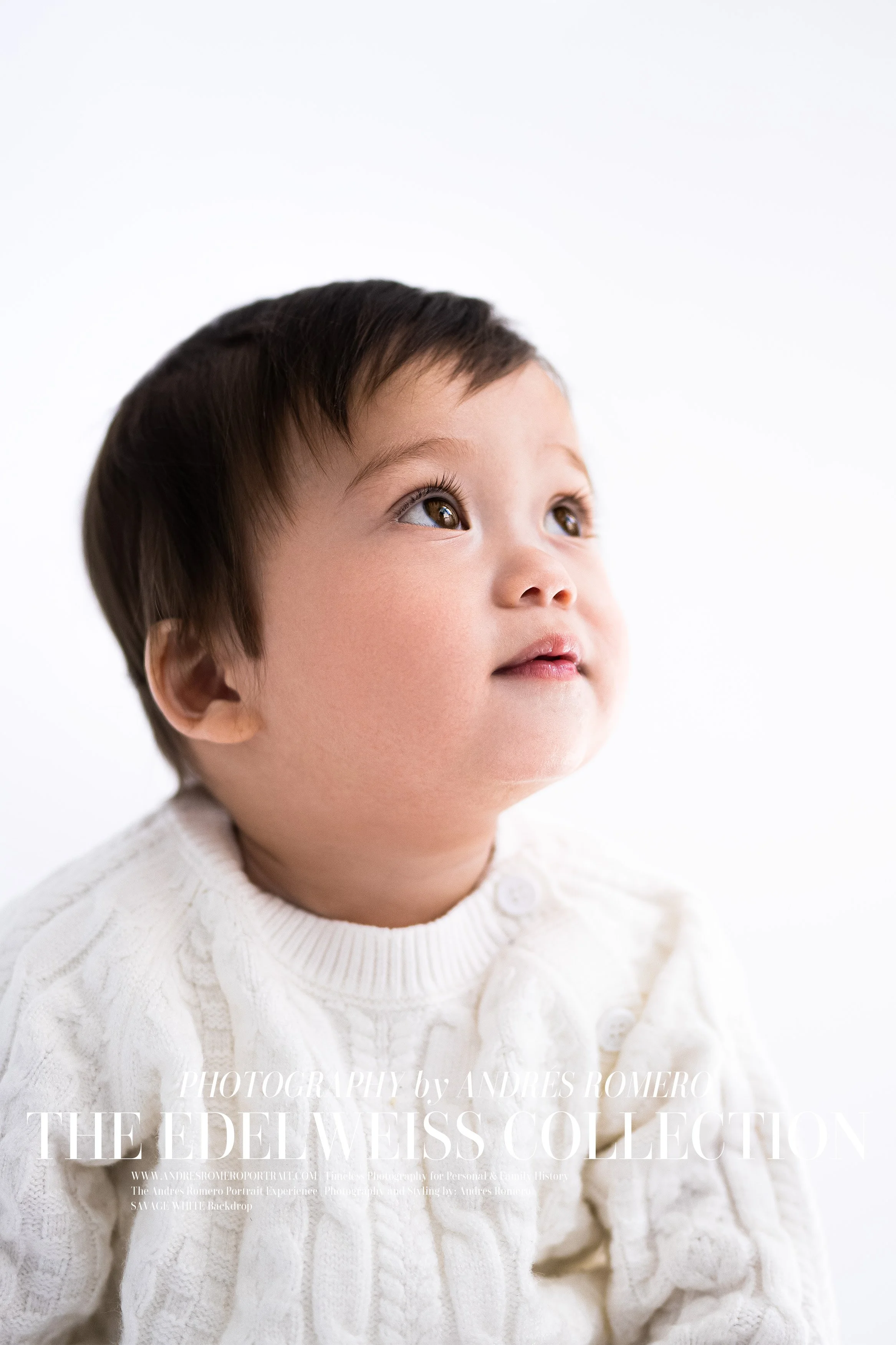 A young child with brown hair and brown eyes wearing a white knitted sweater, looking upward against a plain white background.