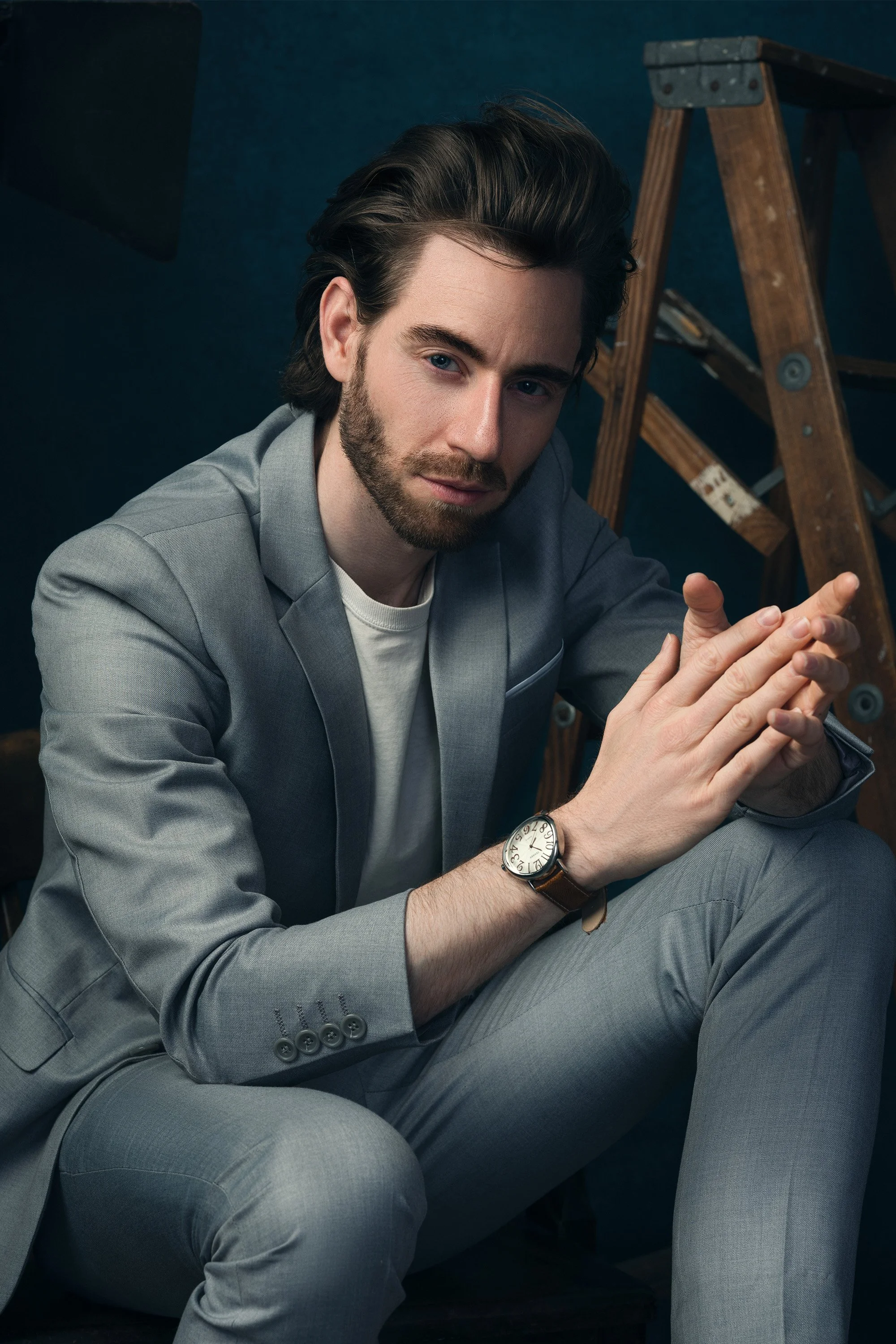 A man with dark hair and a beard, dressed in a light gray suit, sitting on the floor with his hands clasped. Behind him, there is a wooden ladder and a dark, textured wall.