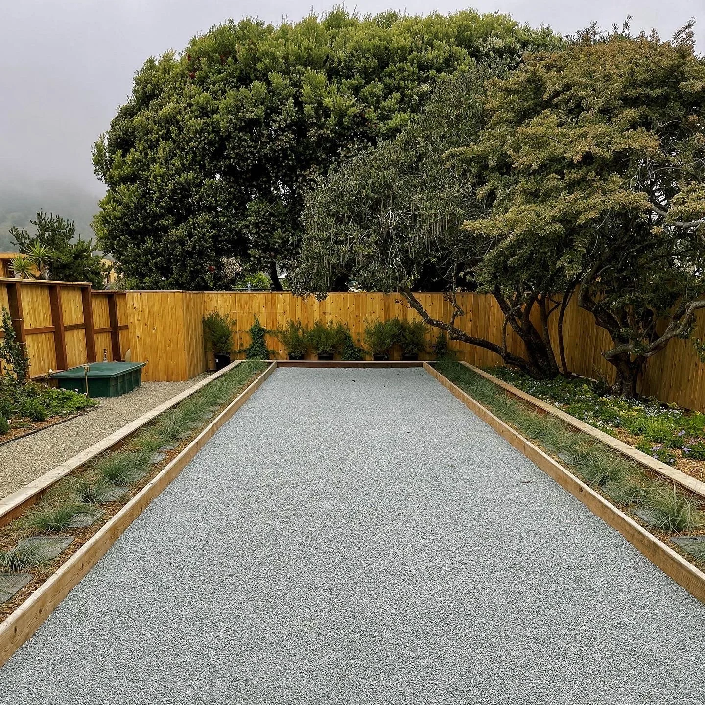 A backyard with a landscaped gravel area, bordered by wooden planks, and lined with plants and small shrubs along a wooden fence. Large trees are visible beyond the fence, and the sky appears overcast.