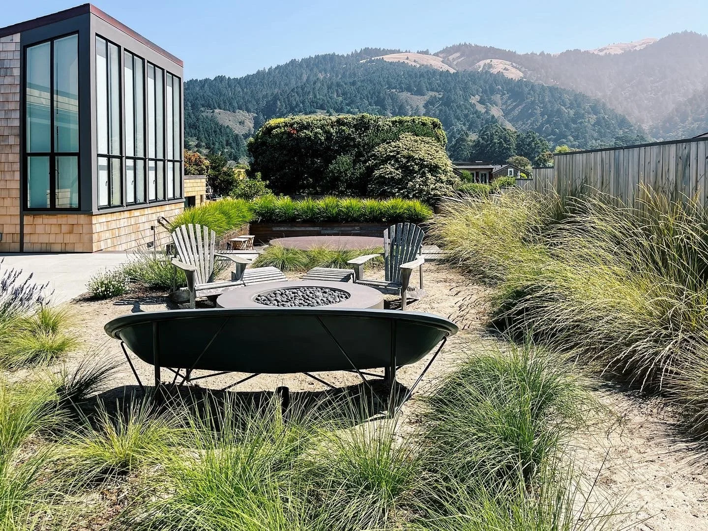 Outdoor patio with Adirondack chairs surrounding a fire pit, chaise lounge, lush grass, and a modern glass house with mountains in the background.