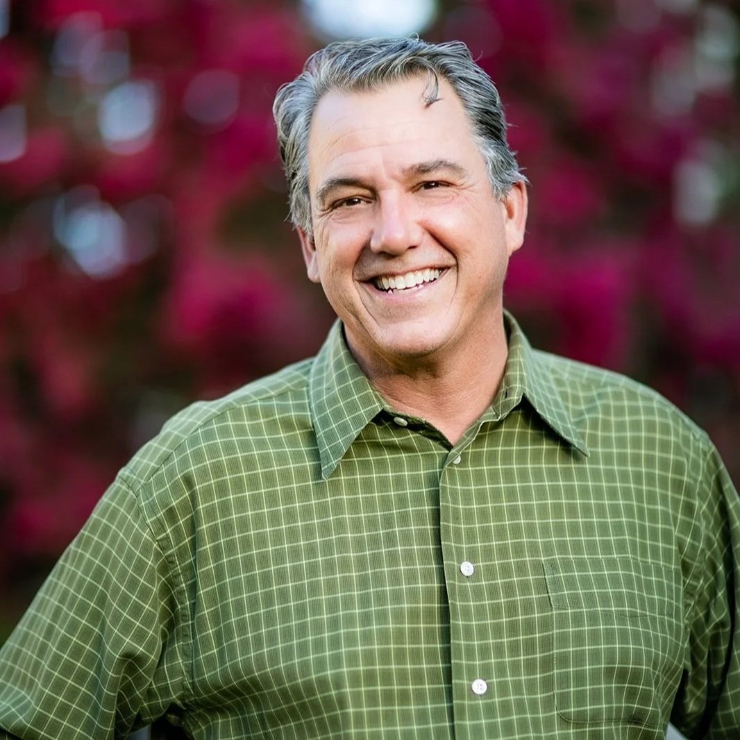 A middle-aged man with gray hair smiling outdoors, wearing a green checkered shirt, with pink and purple flowers blurred in the background.