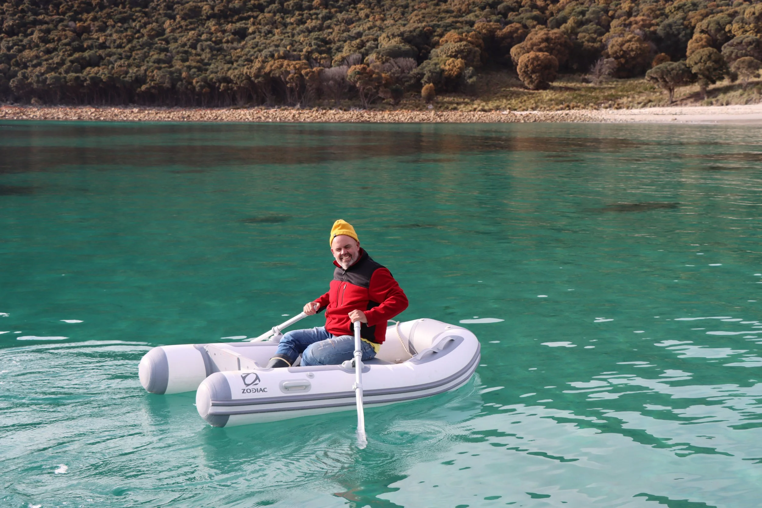 A man with a yellow beanie and red jacket is smiling while sitting in an inflatable boat on clear, greenish water with a forested shoreline in the background.