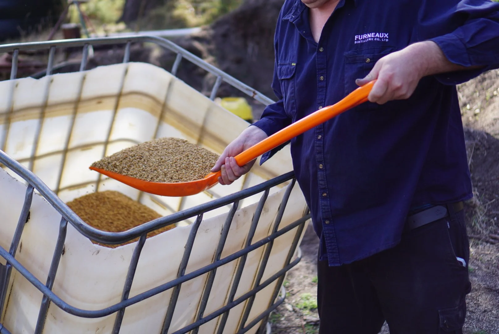 A person wearing a dark blue shirt and black pants is scooping grains from a large white container with an orange scoop, outdoors.