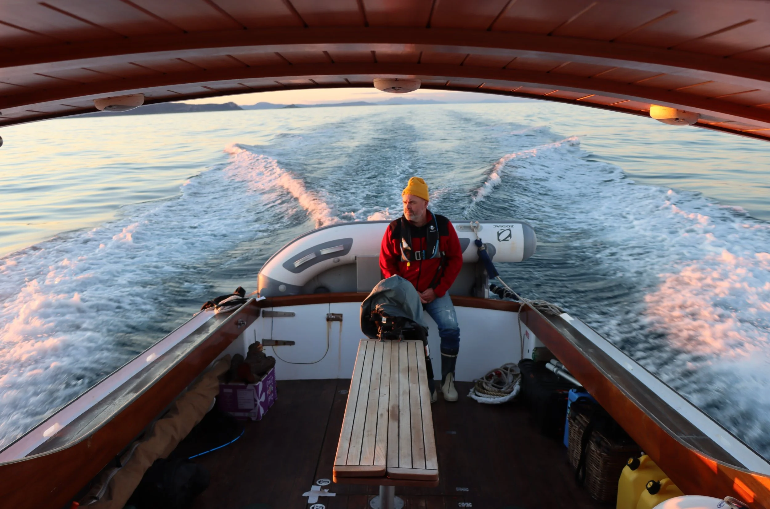 A man with a yellow beanie and red jacket sits at the back of a boat as it moves across calm water during sunset, with a distant shoreline and mountains in the background.