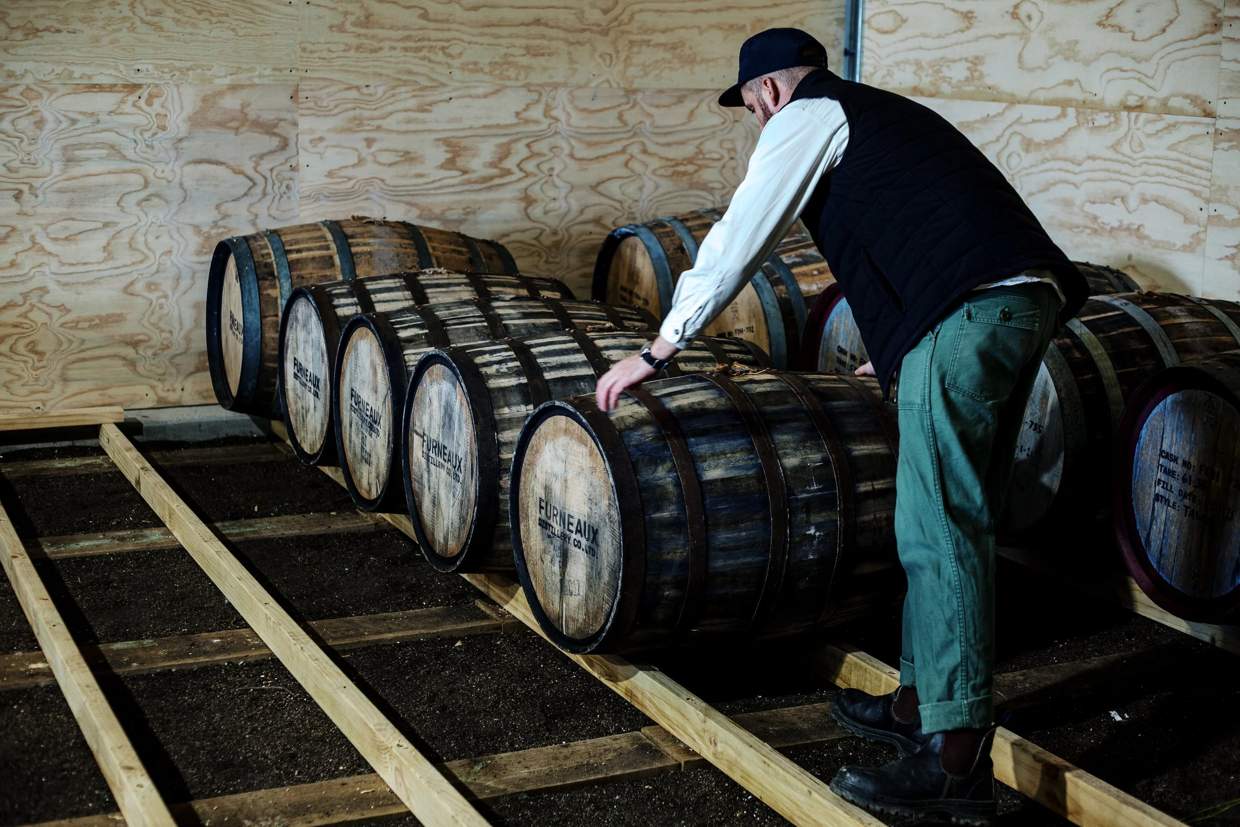 A man in a black vest, white shirt, green pants, and black cap standing over wooden barrels in a warehouse or cellar with unfinished wooden walls.