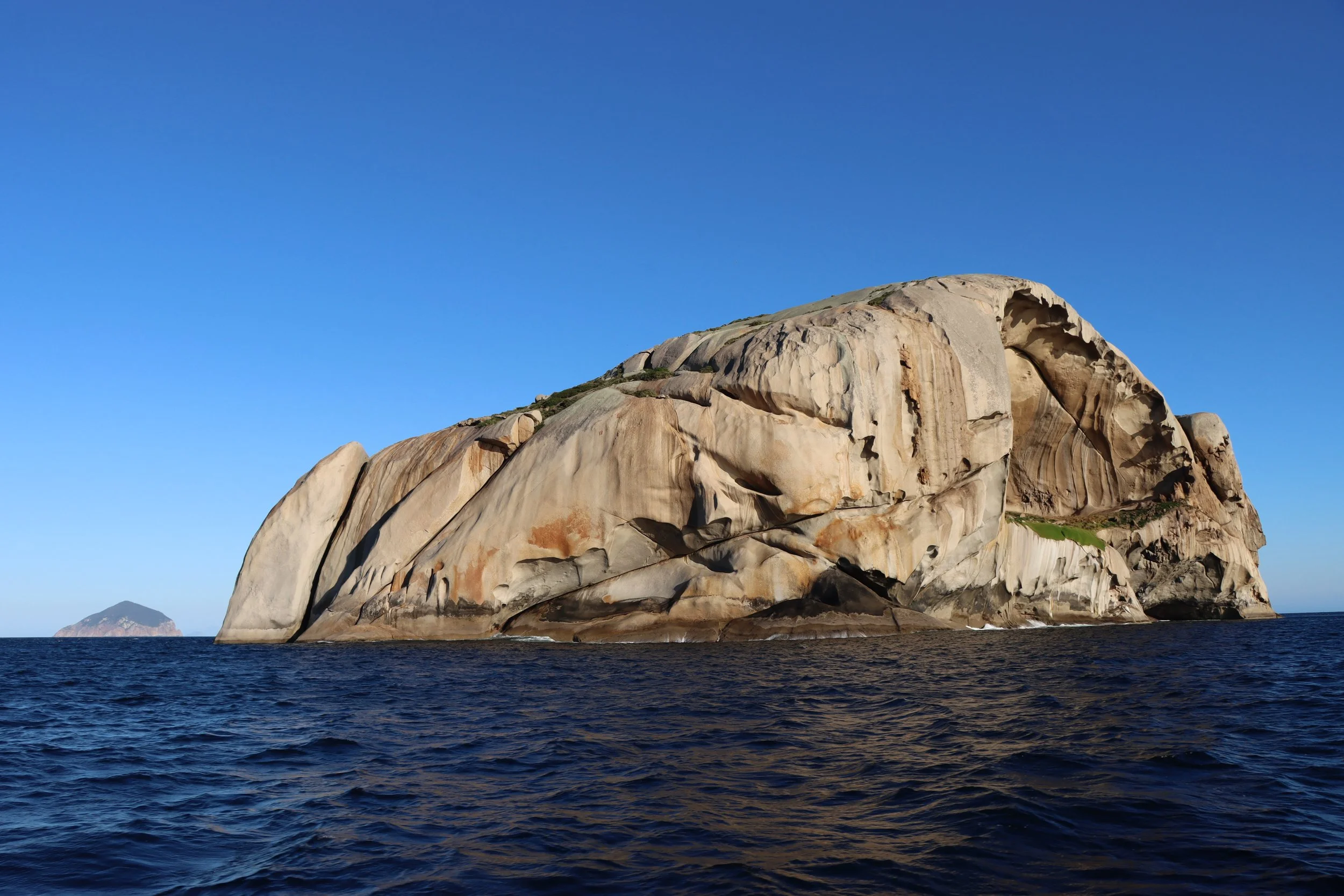 Large granite sea stack rising from the ocean against a clear blue sky, with a small island visible in the background.