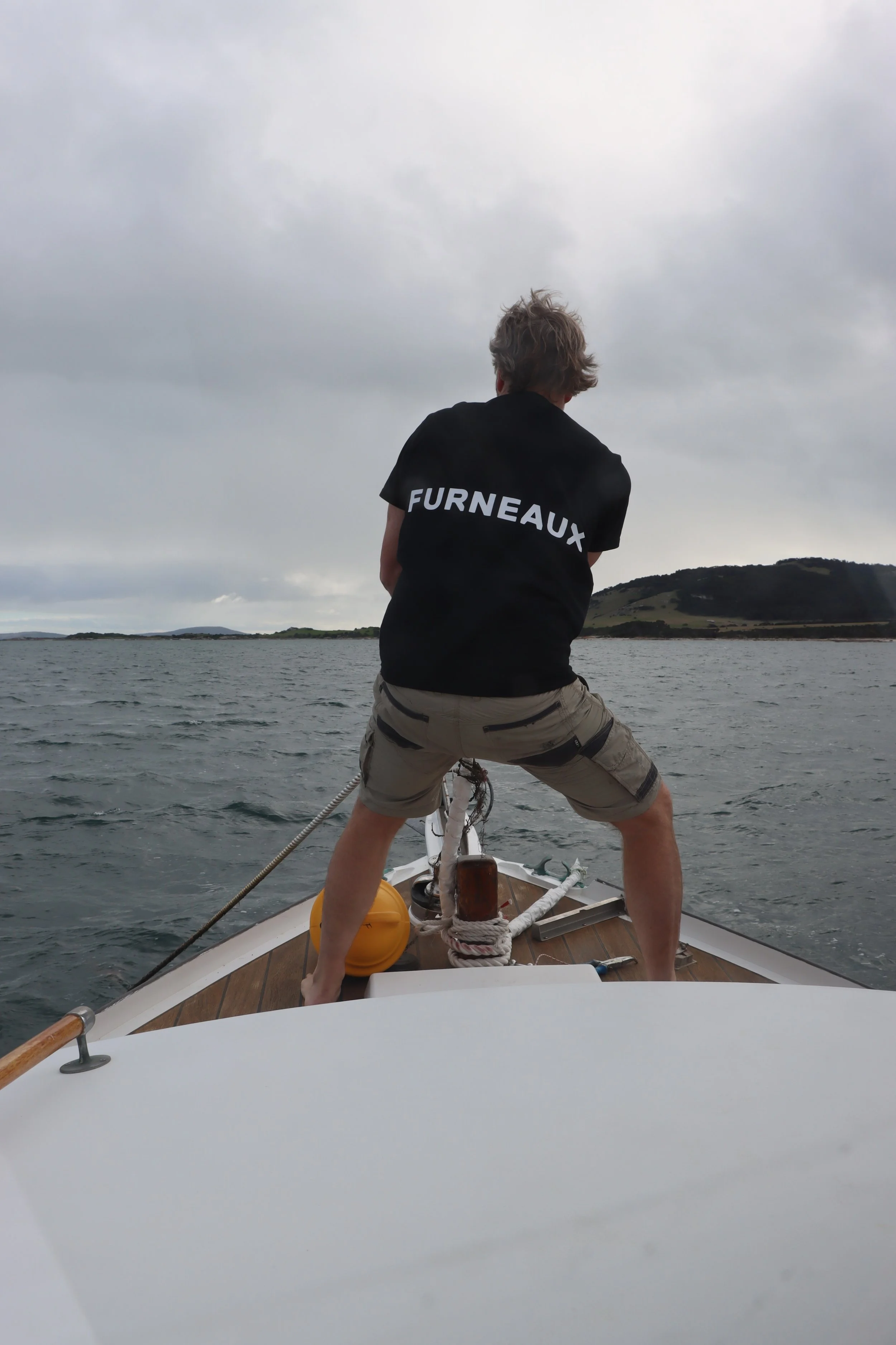 Person standing on the bow of a boat, wearing a black shirt with 'FURNEAUX' printed on the back, holding onto a rope, on a cloudy day over a body of water with land in the distance.