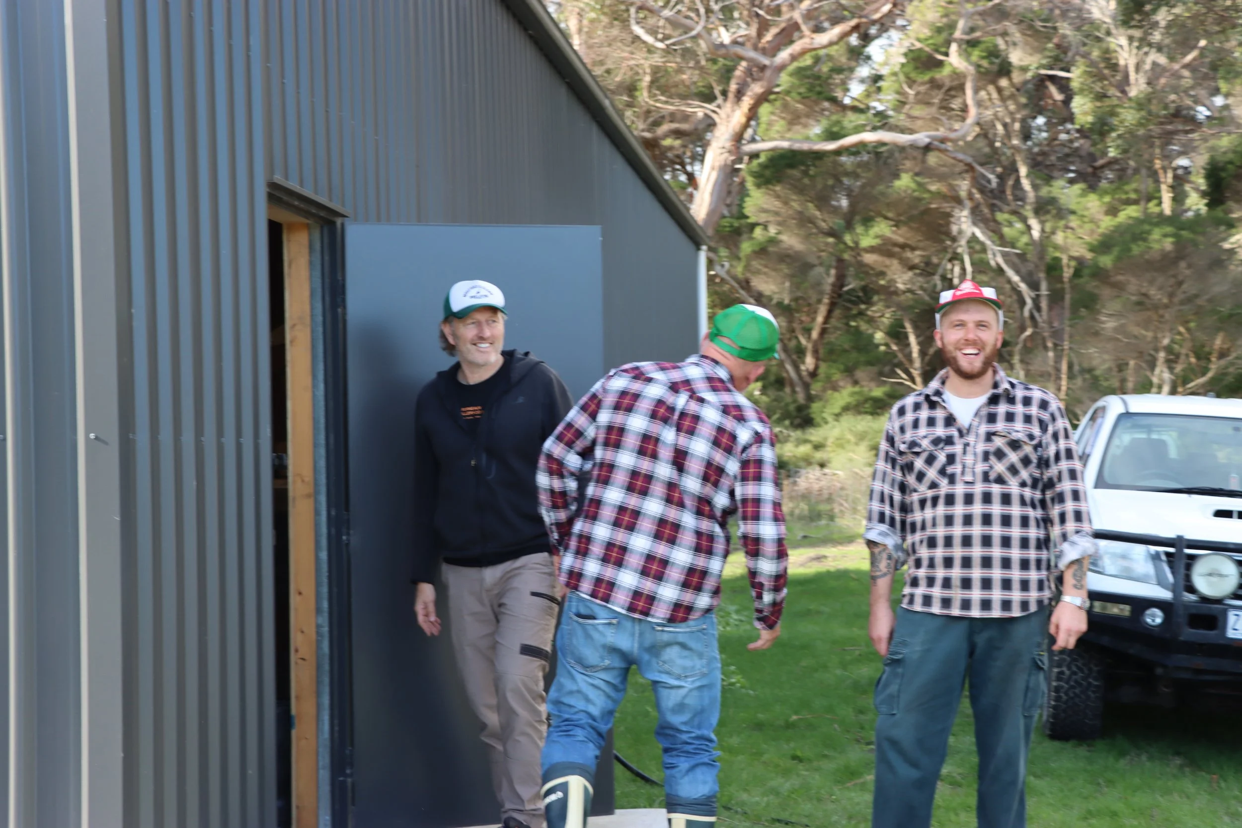 Three men walking outdoors near a metal building, with trees and parked vehicles in the background, one man smiling and wearing a checkered shirt, another in a green cap, and the third in a black hoodie.