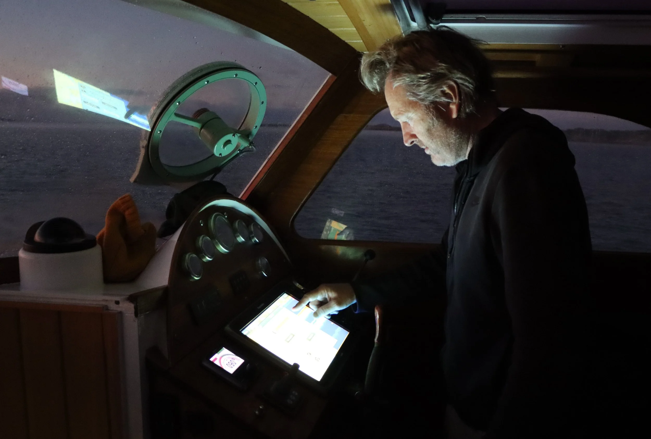 A man with gray hair and beard using a touchscreen device inside a small aircraft cockpit, with a window showing an airplane propeller and dark sky outside.