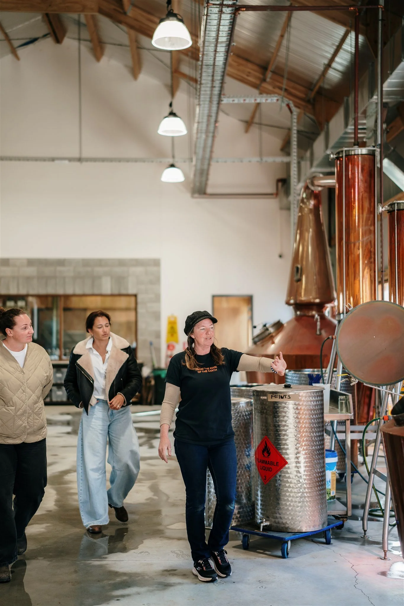 A group of women touring a brewery or distillery, with a guide explaining the brewing process. The setting includes industrial brewing equipment, large copper kettles, and metal tanks with safety warnings.